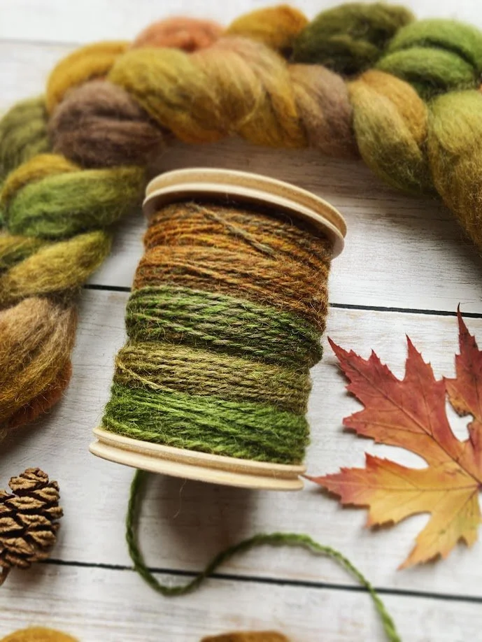 Close-up of a spool of multicolored yarn in shades of green, brown, and orange, with a braided multicolored yarn strand nearby, set on a white wooden surface with a red maple leaf and pine cone.