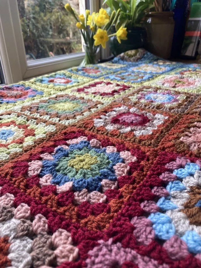 Colorful crocheted blanket with various square patterns draped over a surface near a window with yellow daffodils in the background.