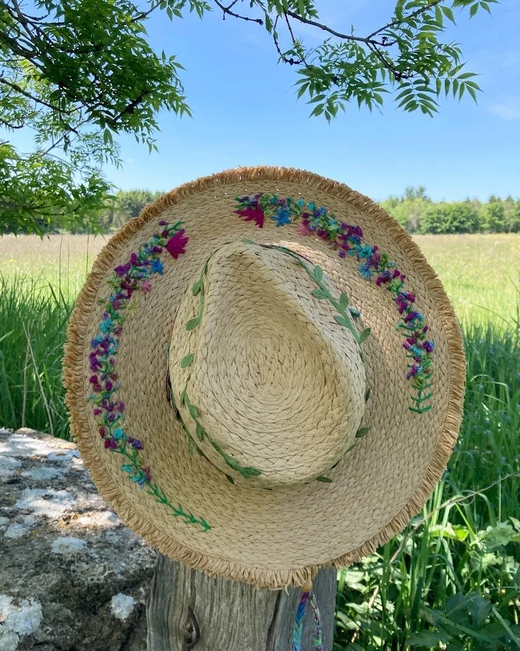 A woven straw hat with colorful floral embroidery hanging on a wooden post in a grassy outdoor area under a clear blue sky.