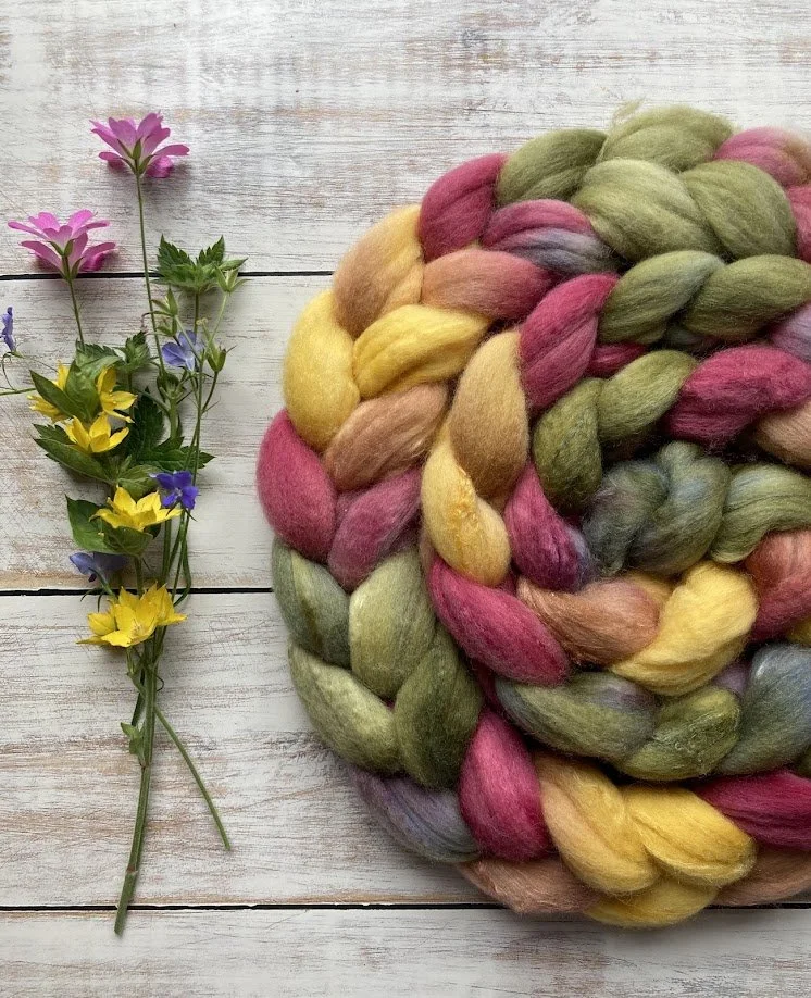 Colorful braided wool surrounded by small pink, purple, and yellow flowers on a white wooden surface.
