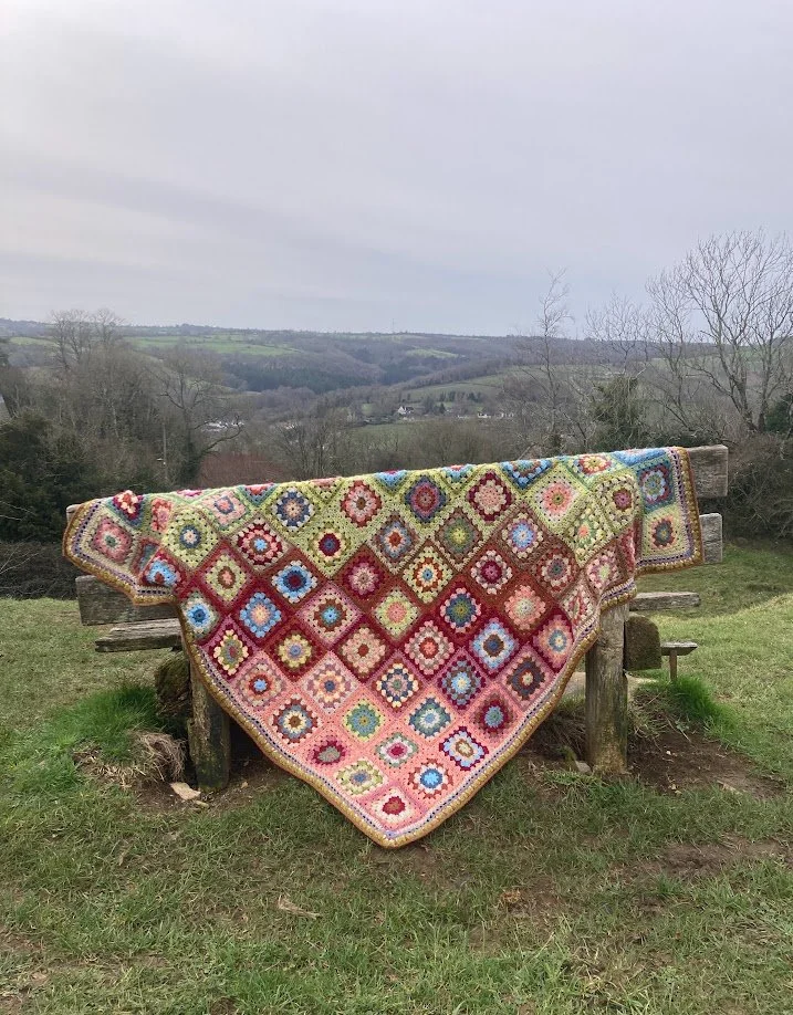 Colorful crochet granny square blanket draped over a wooden bench outdoors with a scenic countryside background.