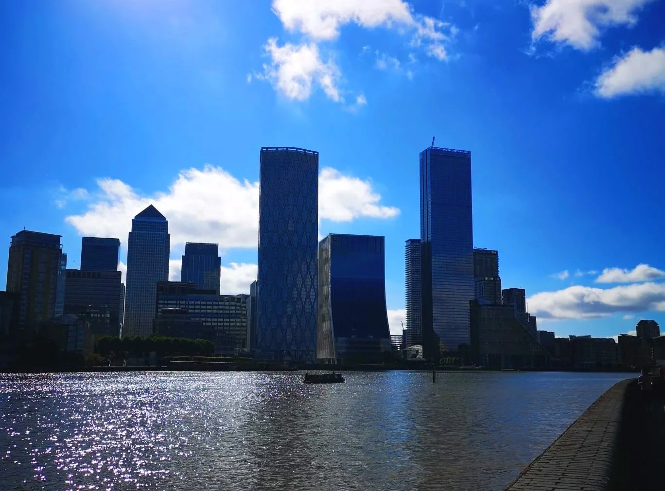 City skyline with tall modern skyscrapers along a river under a blue sky with clouds.