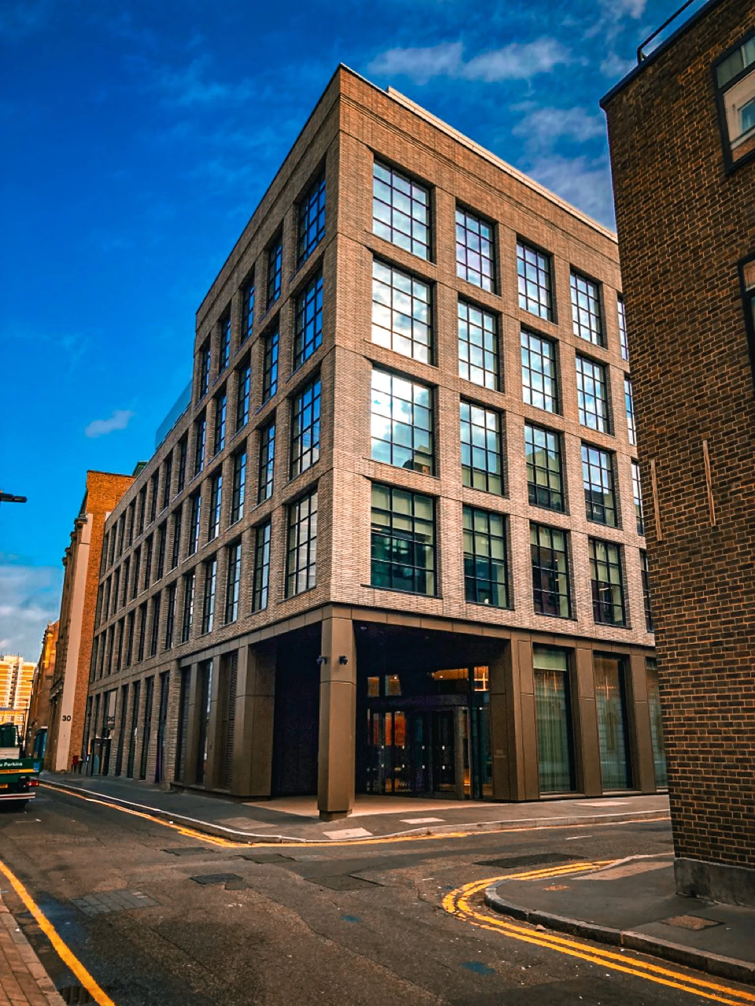A modern multi-story brick building with large glass windows, situated on a city street corner beneath a blue sky with some clouds.