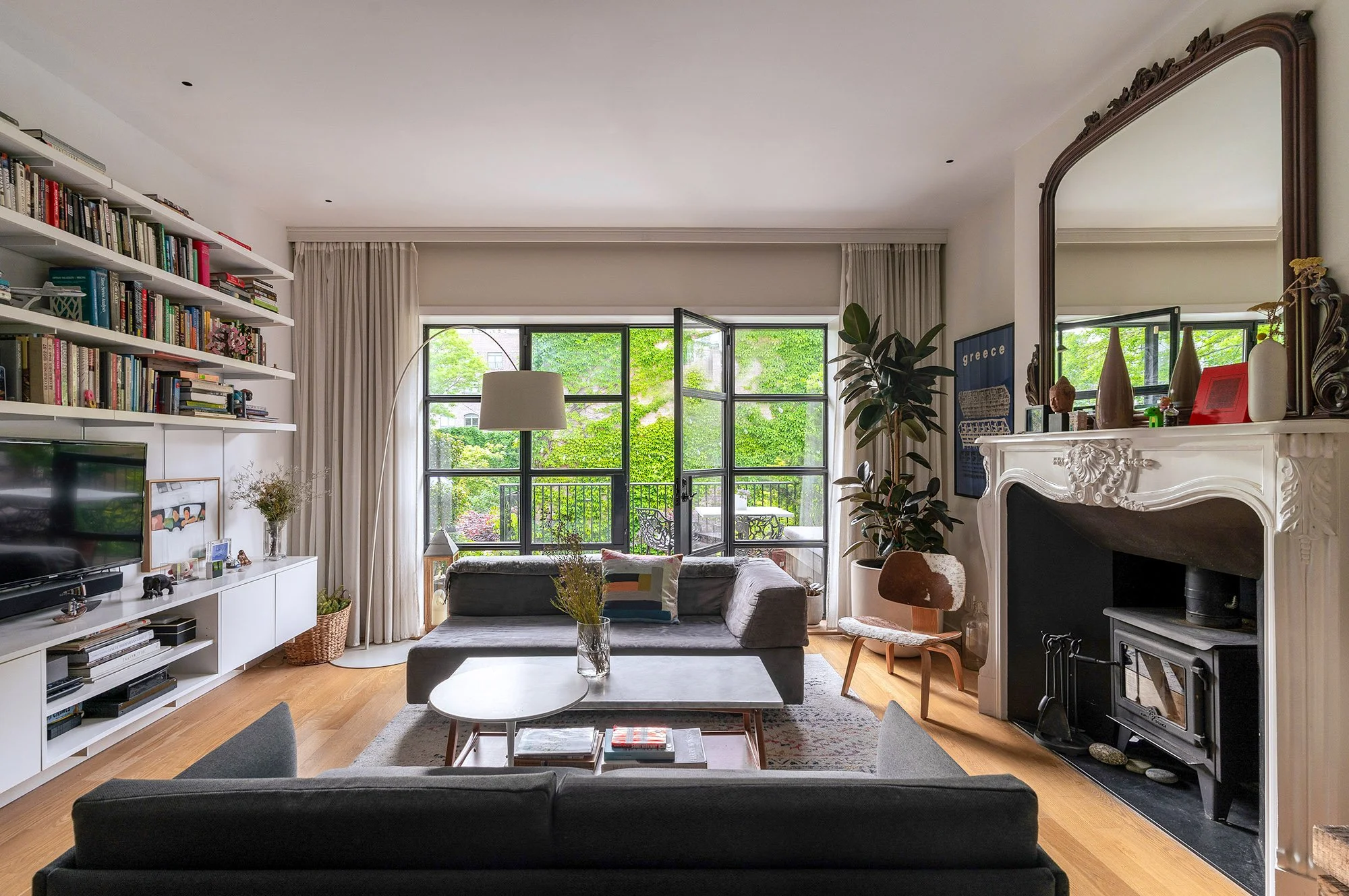 Contemporary living room with grey sofas, a large window with curtains, bookshelves filled with books, a flat-screen TV, and a decorative fireplace with a large mirror. There's a side chair, houseplants, and a coffee table with magazines. A wooden floor and a view of greenery outside are visible.