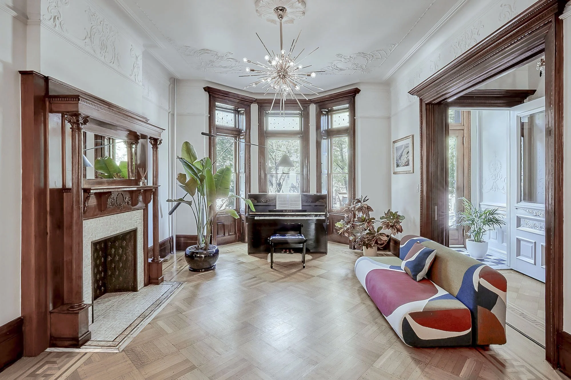 Elegant living room with a colorful modern sofa, upright piano, large indoor plants, ornate fireplace, and a decorative chandelier.