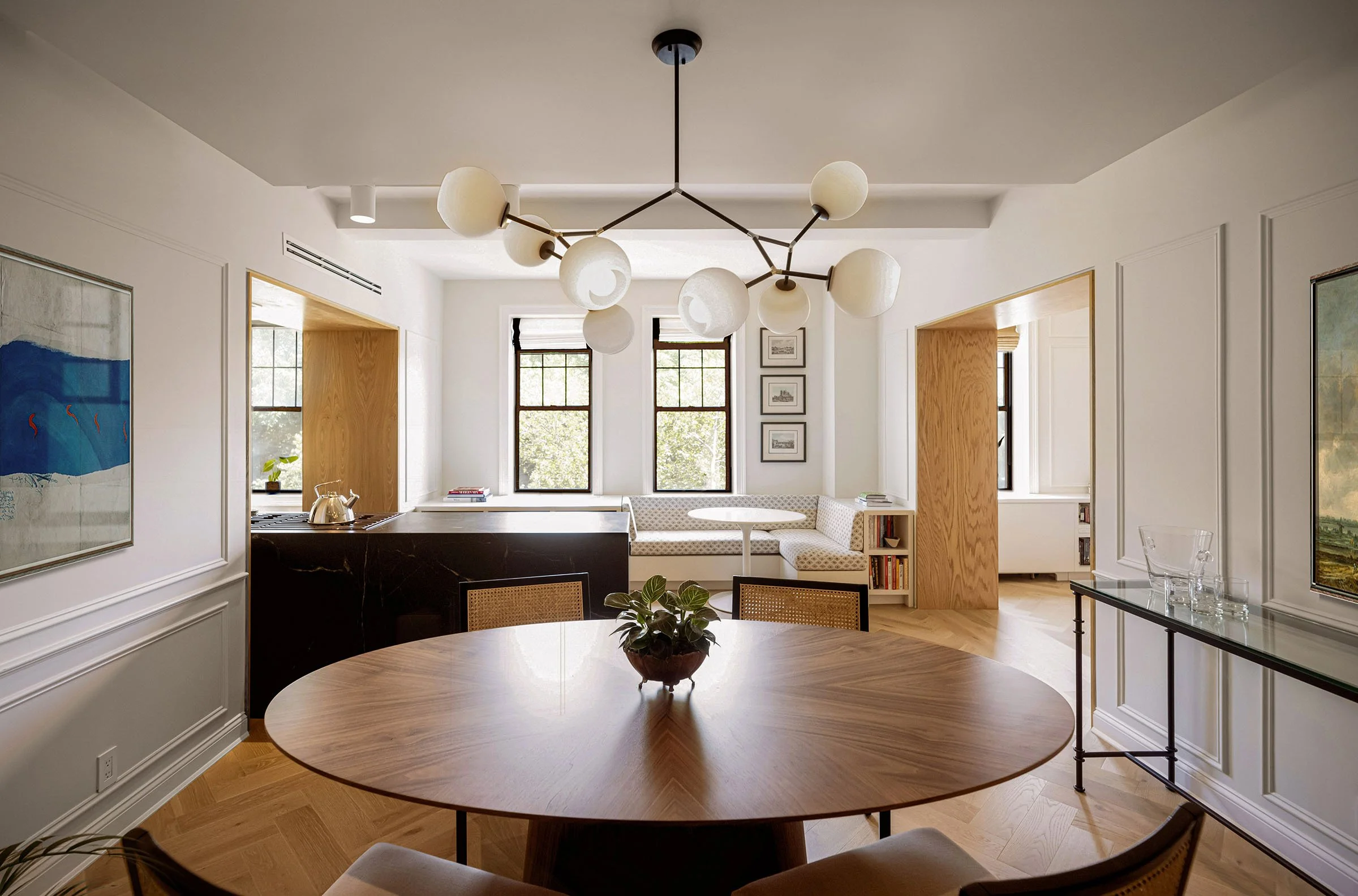 Modern, stylish dining room with a round wooden table and chairs, a potted plant centerpiece, large chandelier, and wall art. The room includes a window nook with a small round table, cushioned seating, and windows overlooking greenery. Light wood floors and neutral-toned walls complement the sleek decor.