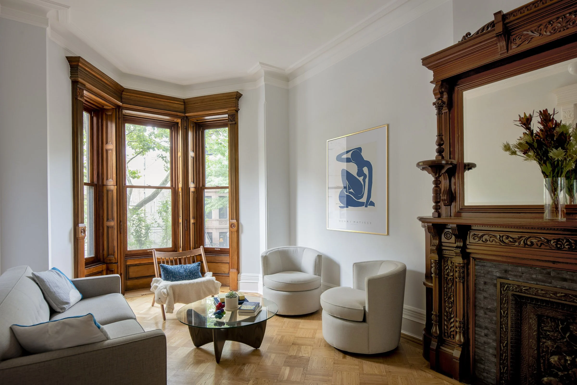 A stylish living room with parquet flooring, featuring a gray sofa, a glass coffee table, two round chairs, and a wooden armchair by bay windows. The room has a decorative wooden fireplace and a modern art print on the wall.