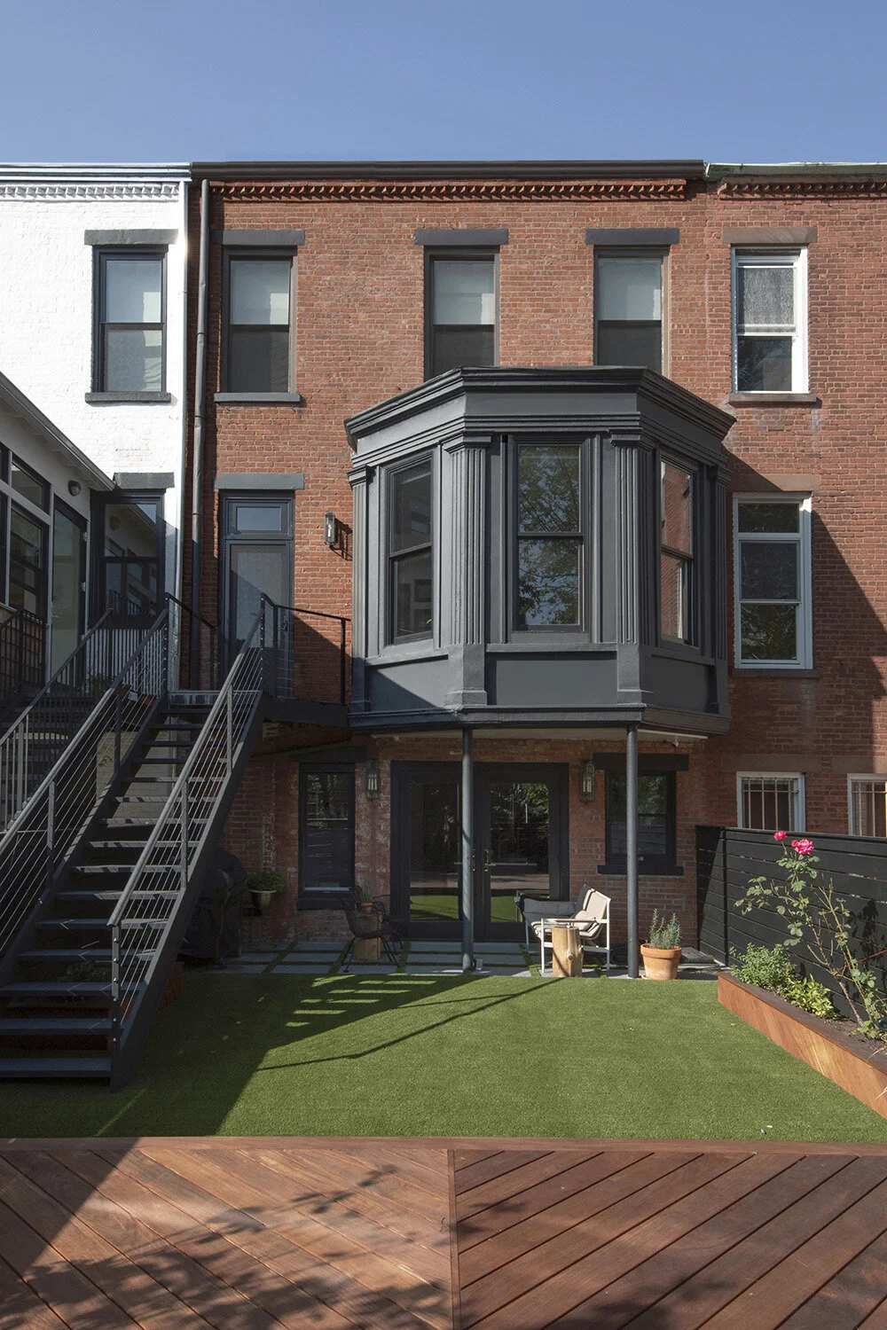 Backyard view of a three-story brick townhouse with a modern glass and black framed bay window, metal exterior staircase, glass door entry, artificial grass lawn, wooden deck, and outdoor seating area.