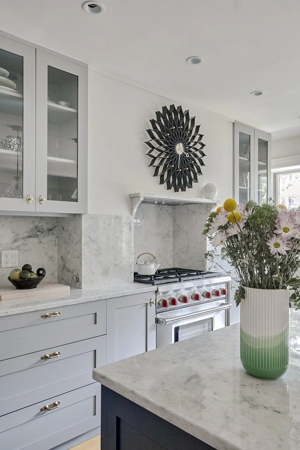 Modern kitchen with gray cabinets, marble countertops, a stainless steel range with red knobs, a decorative wall clock, and a vase of flowers on the island.