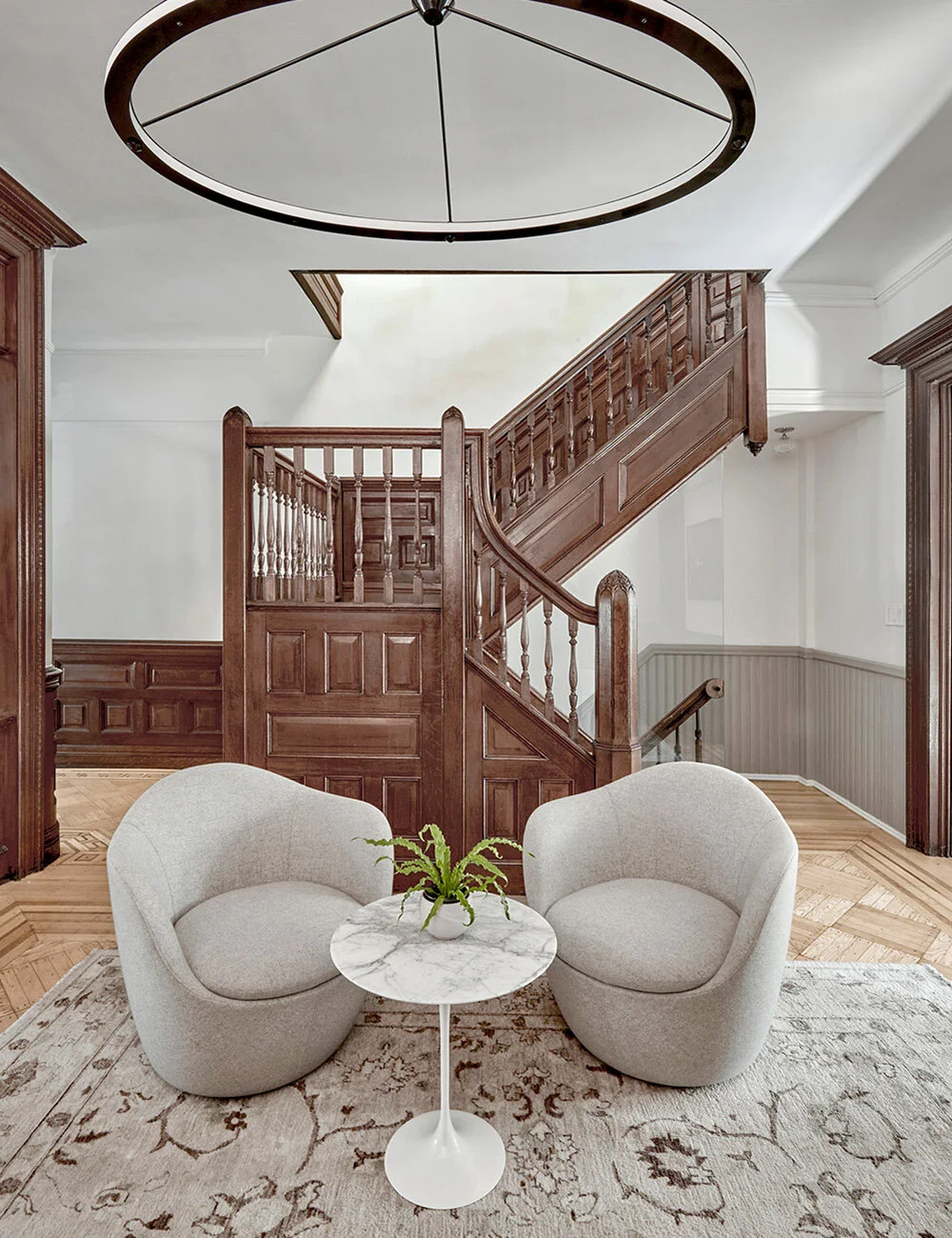 Two gray armchairs with a small round marble table in between, placed on a patterned rug, in front of a wooden staircase with a railing, and a circular light fixture above.