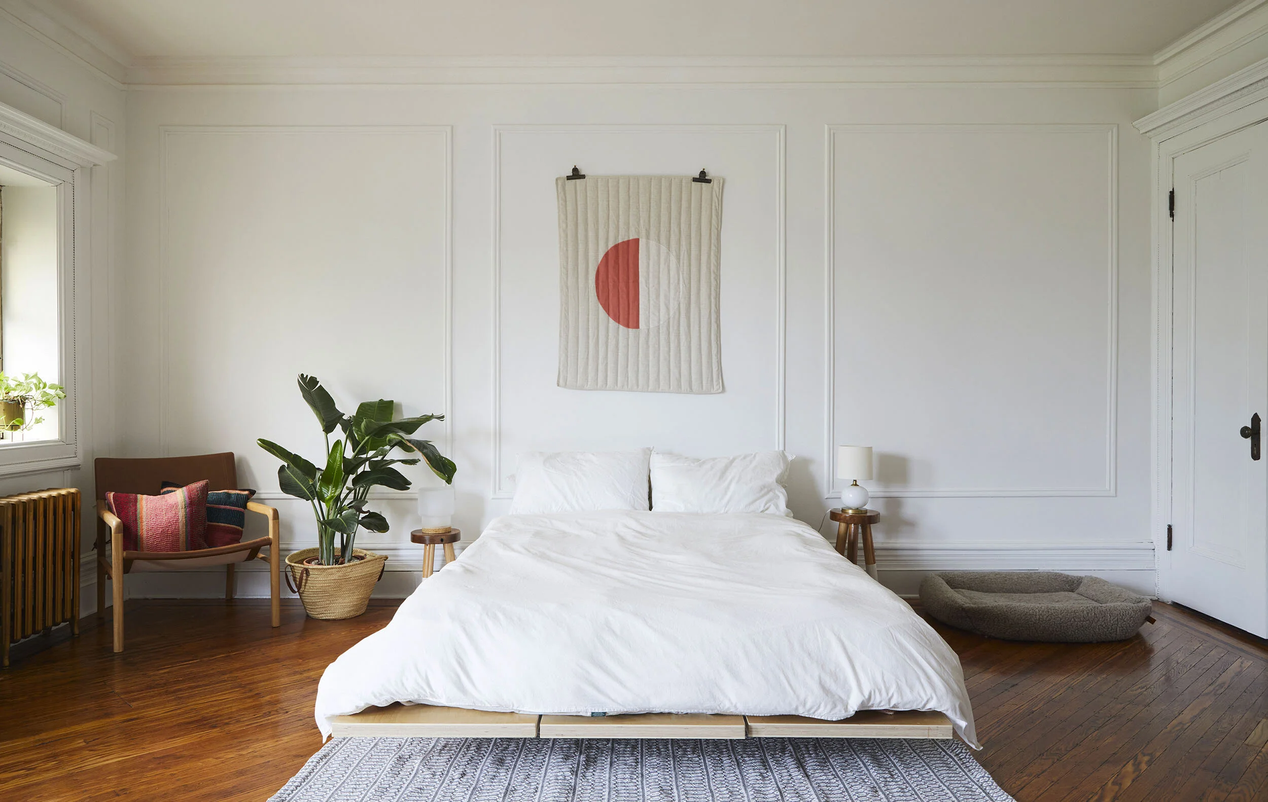 Minimalist bedroom with white bedding, wooden floor, and wall art. A plant and chair with colorful pillows sit next to the bed. Two nightstands with lamps are on either side, and a pet bed is on the floor.