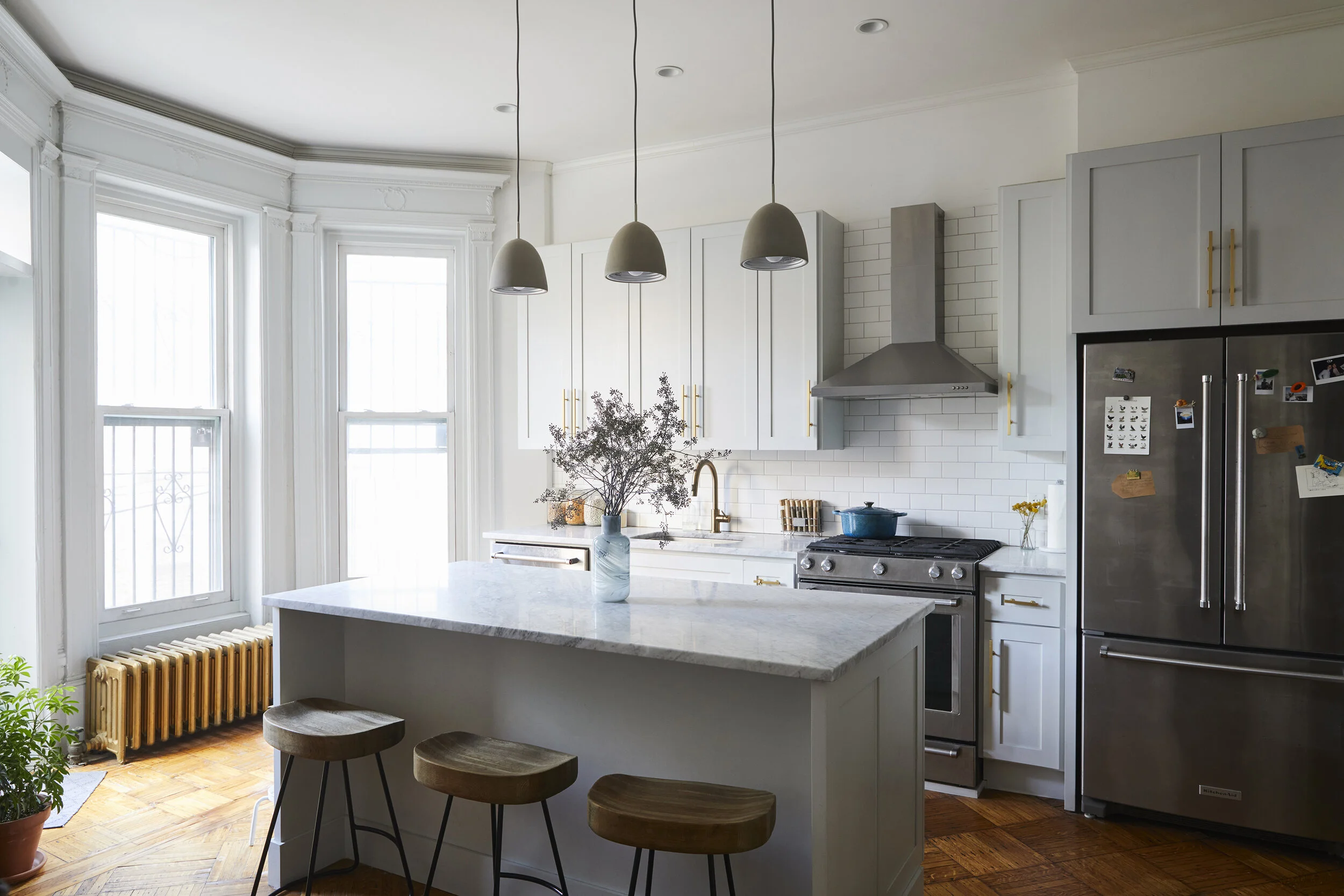 Modern kitchen with white cabinets, marble countertops, stainless steel appliances, wooden stools, pendant lights, and a vase with flowers on the island.