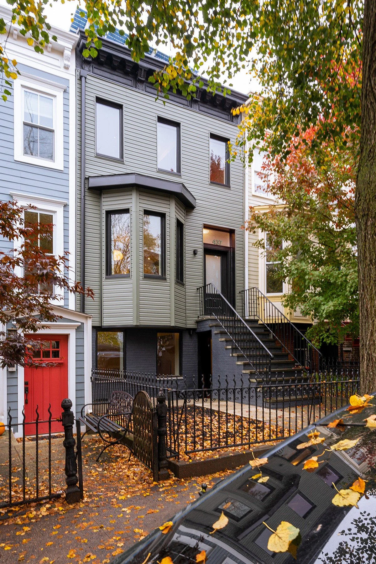 Victorian-style townhouse with green and gray exterior, black railings, red door, surrounded by autumn leaves and trees.