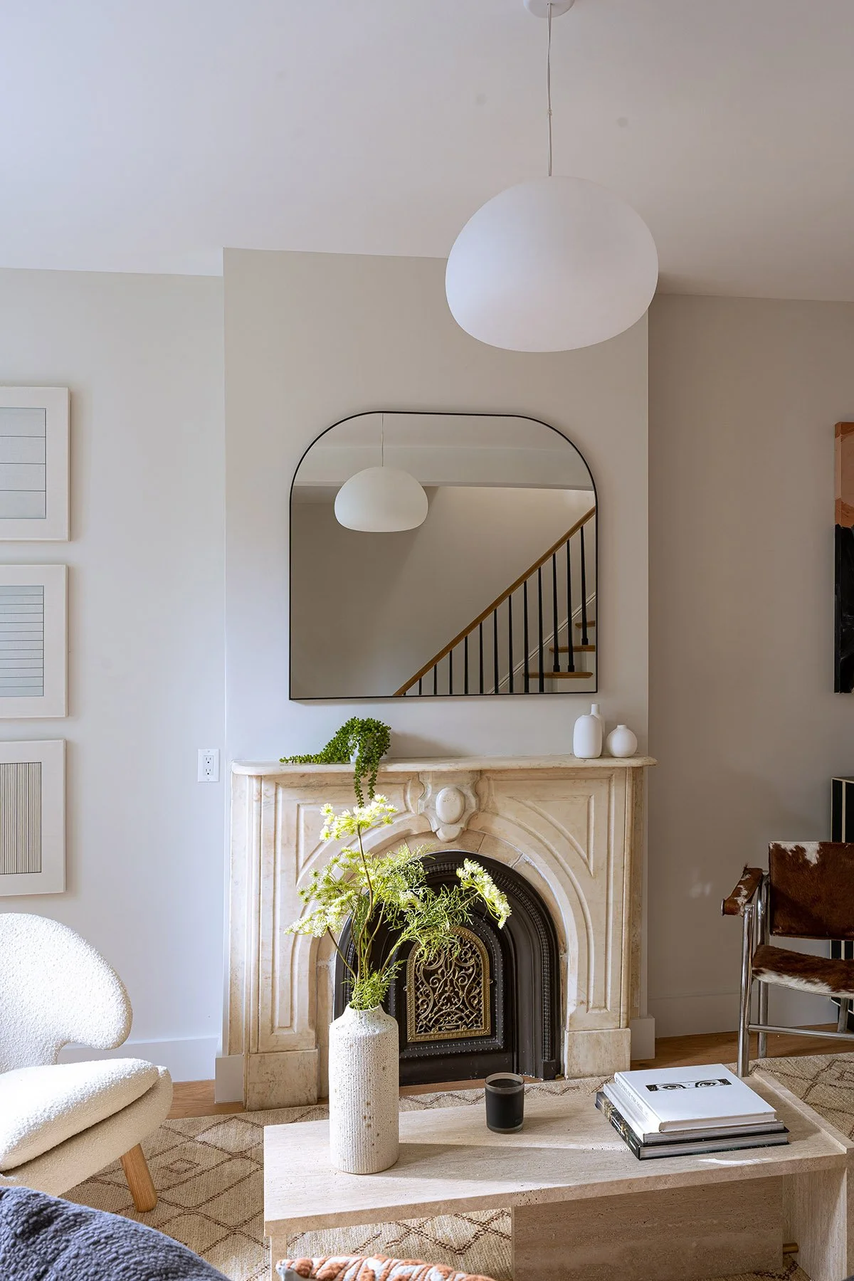 Modern living room with a marble fireplace, large mirror, white pendant light, beige armchair, and a coffee table with vase and books.