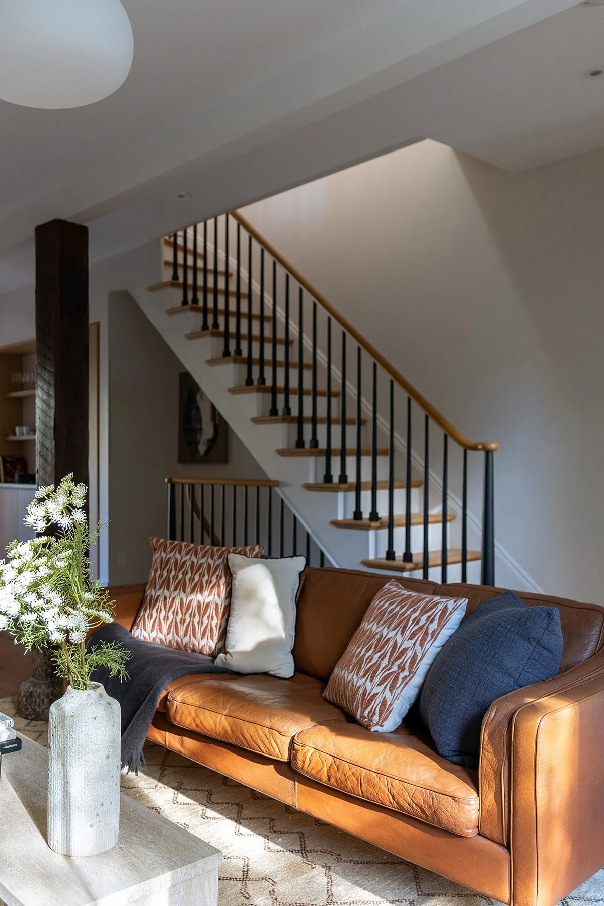 Modern living room with a brown leather sofa, decorative pillows, and a staircase in the background. A white vase with flowers is on the coffee table.