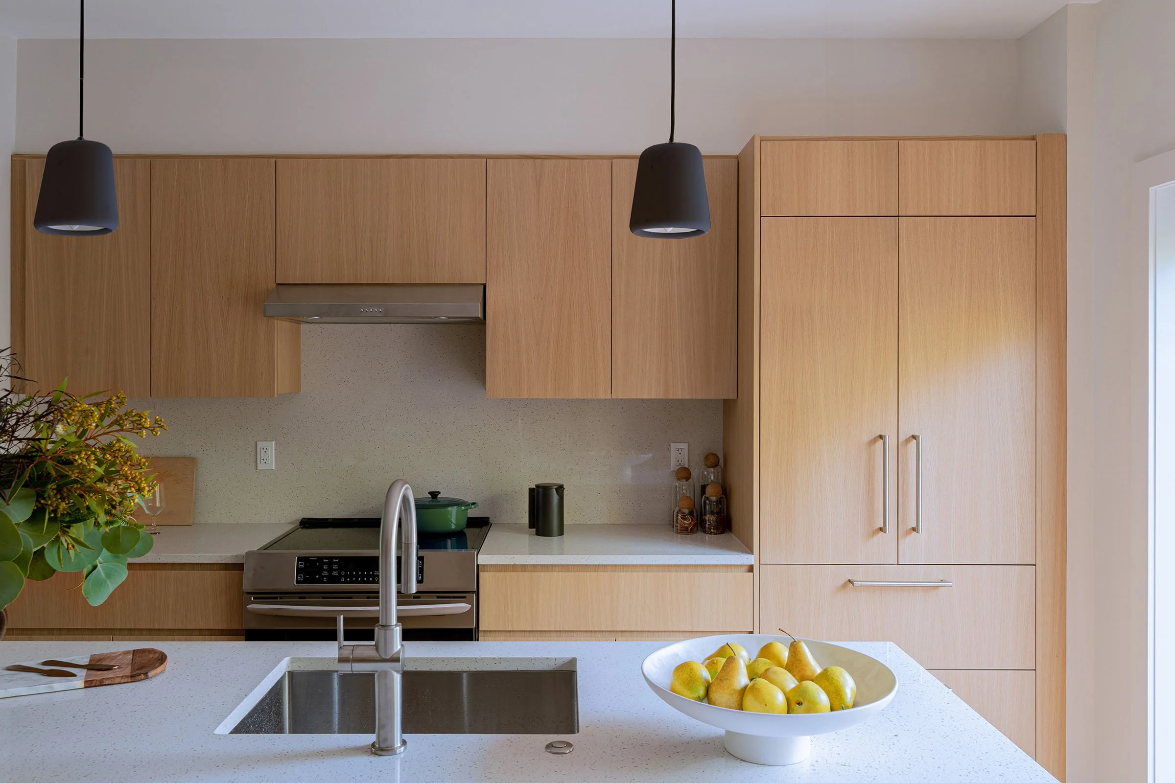 Modern kitchen with light wood cabinets, white countertops, a stainless steel sink, a stovetop, and a bowl of pears on the island.