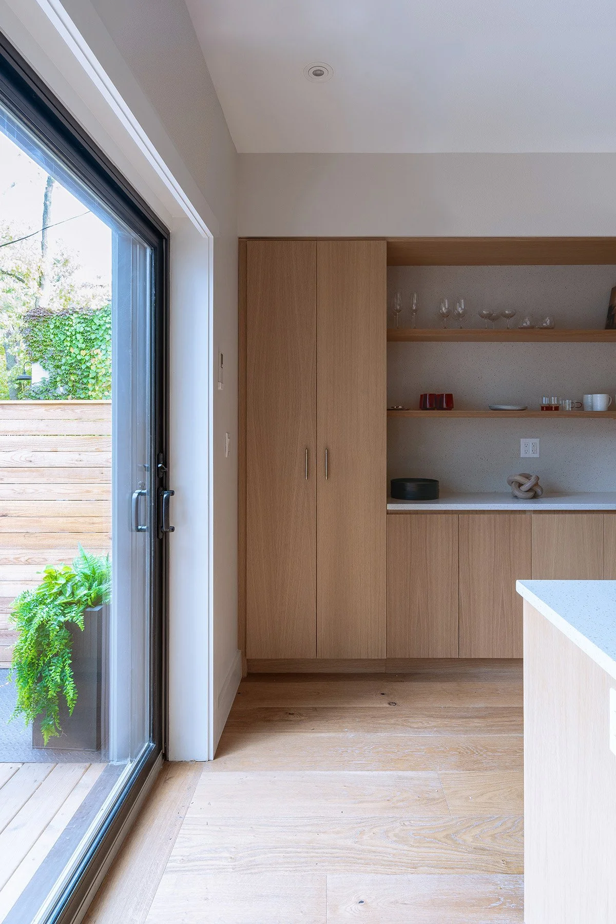 Modern kitchen interior with wooden cabinets and shelves, glassware, and red cups. A large sliding glass door opens to an outdoor deck with greenery. Light wood flooring and white countertops are visible. Minimalist and clean design.