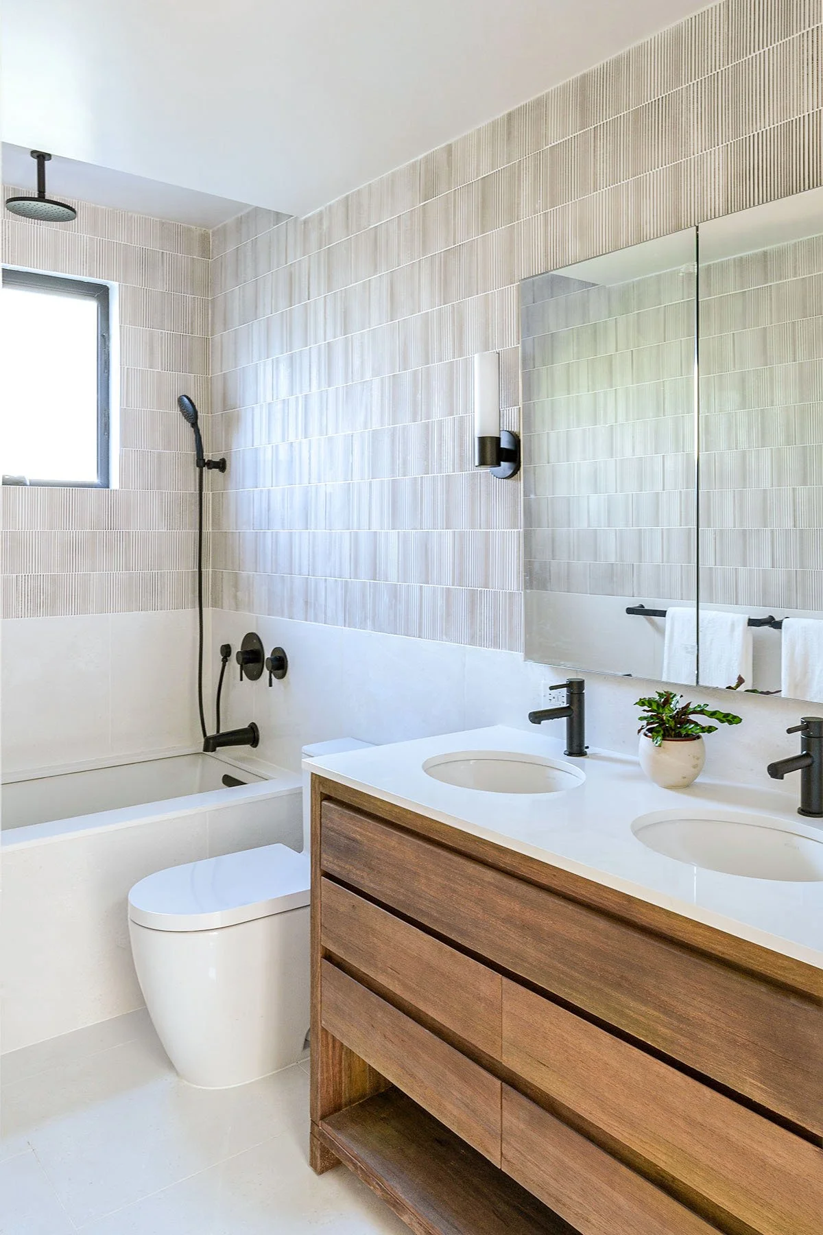 Modern bathroom with a double vanity, wooden cabinet, and black fixtures. Features include a bathtub with a window above, light-colored wall tiles, a round mirror, and a small potted plant on the countertop.