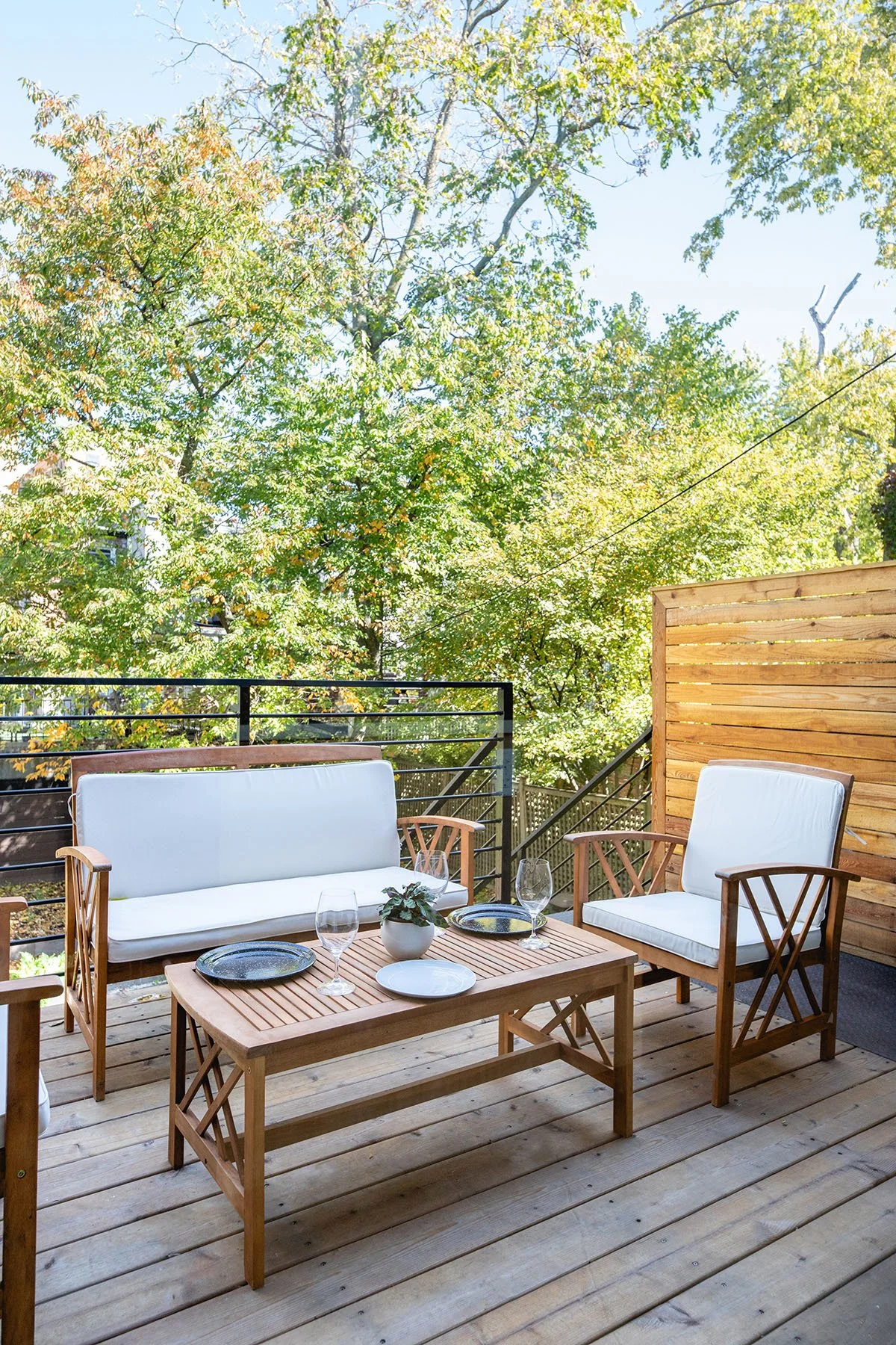 Patio with wooden furniture, including a sofa and chairs with white cushions, a coffee table with plates and glasses, surrounded by trees and greenery.