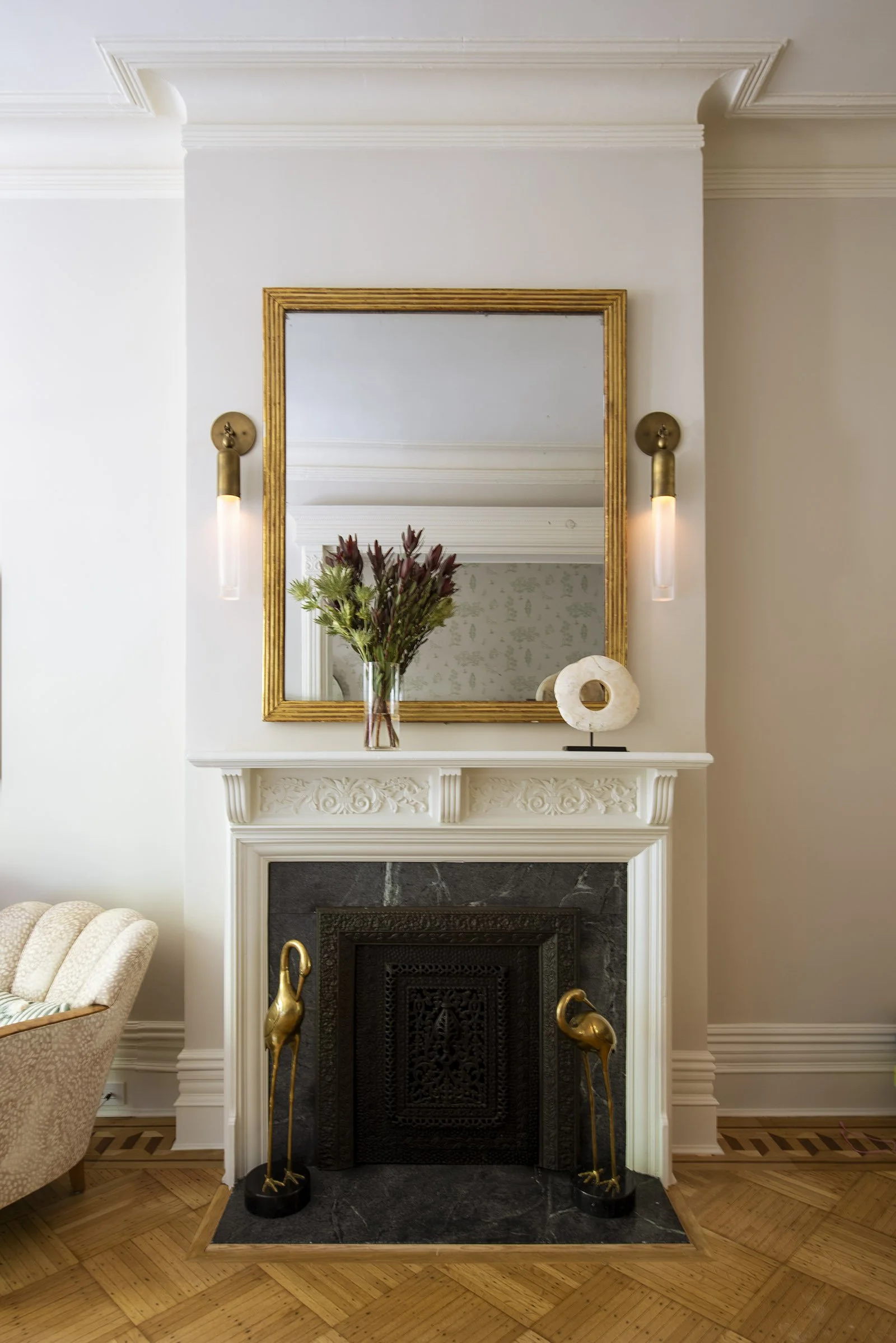 Elegant fireplace with decorative white mantel, gold-framed mirror, vase of flowers, and artistic sculpture; flanked by two wall sconces and gold bird statues on the hearth. at a Park Slope Brooklyn brownstone project