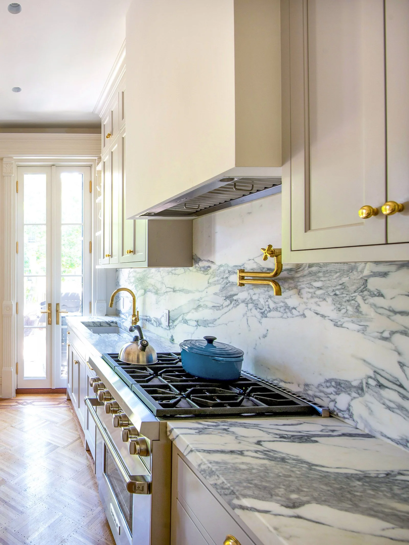 Modern kitchen with marble countertops, cream cabinets, gold hardware, gas stove with blue pot, and glass doors.A jewel-box renovation of a 3-story historic Brooklyn Brownstone aimed at bringing all the modern functionalities to a growing family