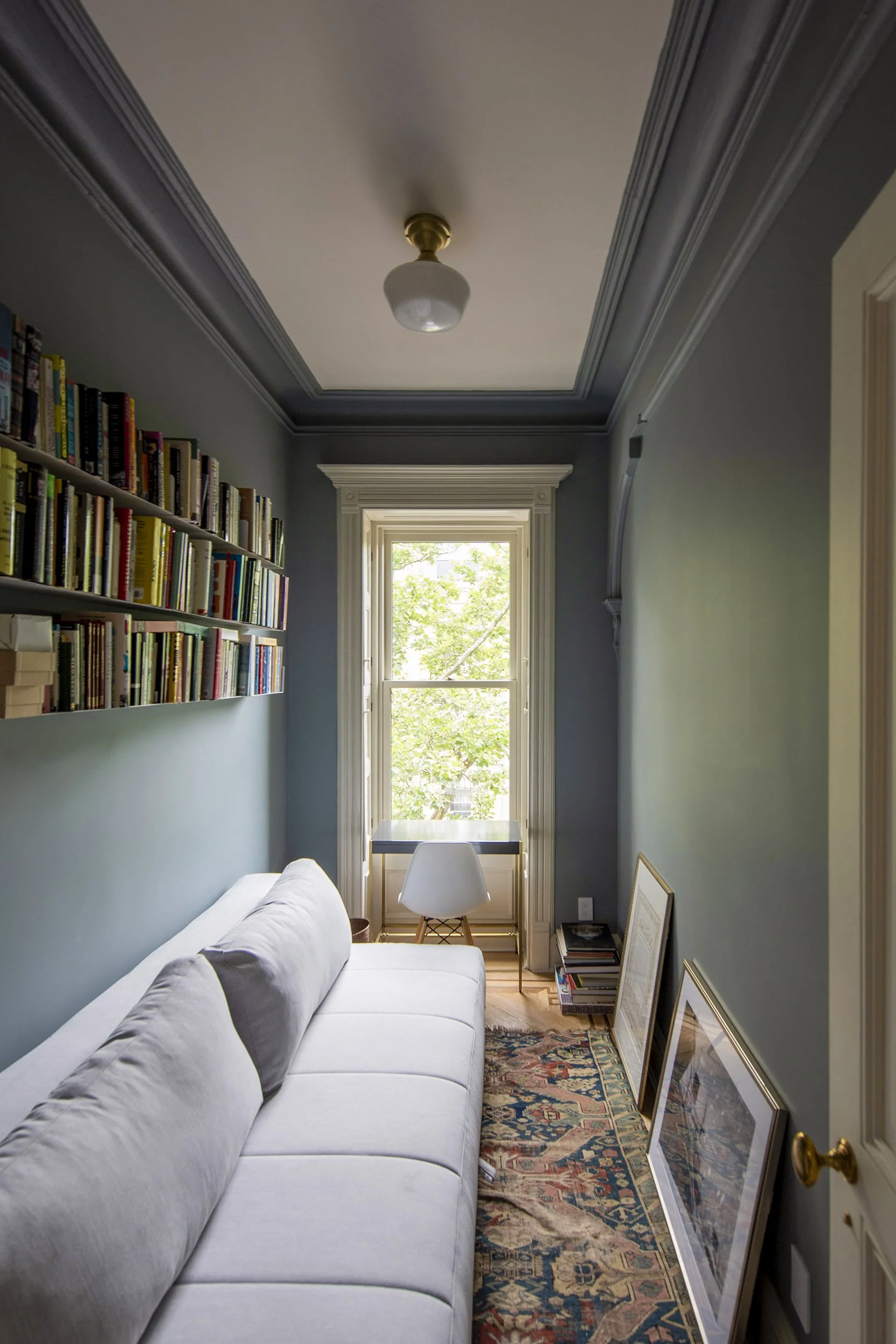 Cozy narrow room with a white sofa, bookshelf, window, rug, and framed pictures. at a Park Slope Brooklyn brownstone project