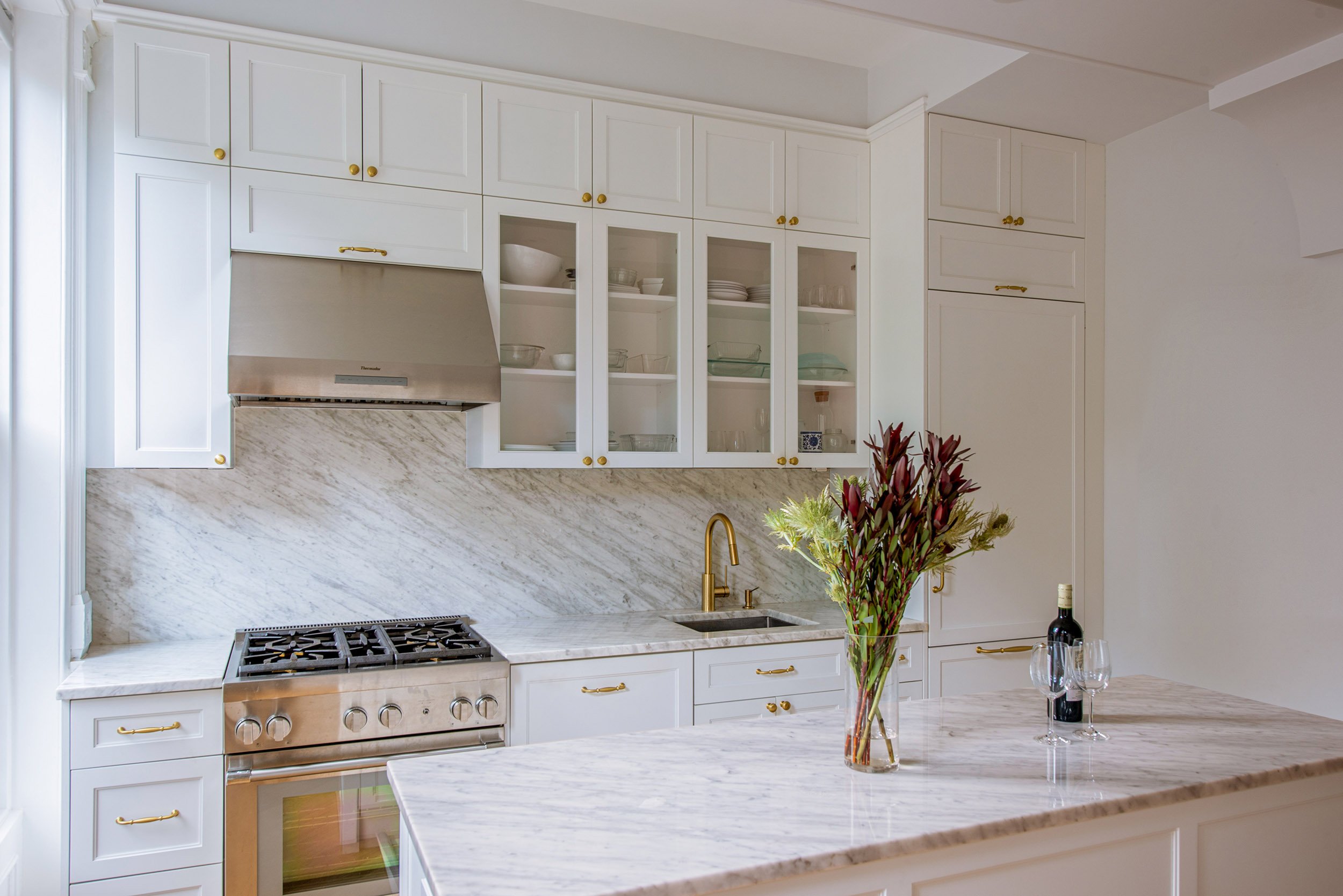 Modern kitchen with white cabinetry, marble countertops, gas stove, and stainless steel vent hood; vase of flowers and wine bottle on the island.