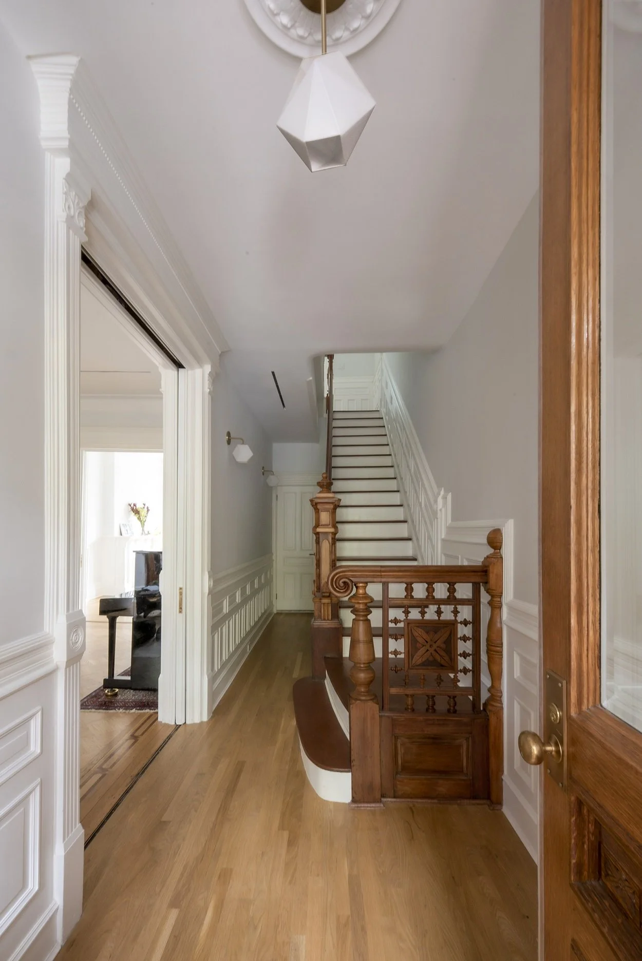 Interior of a classic home with a wooden staircase, ornate railing, and decorative molding. A geometric pendant light hangs from the ceiling above the hardwood floor. An adjacent room with a fireplace is partially visible.