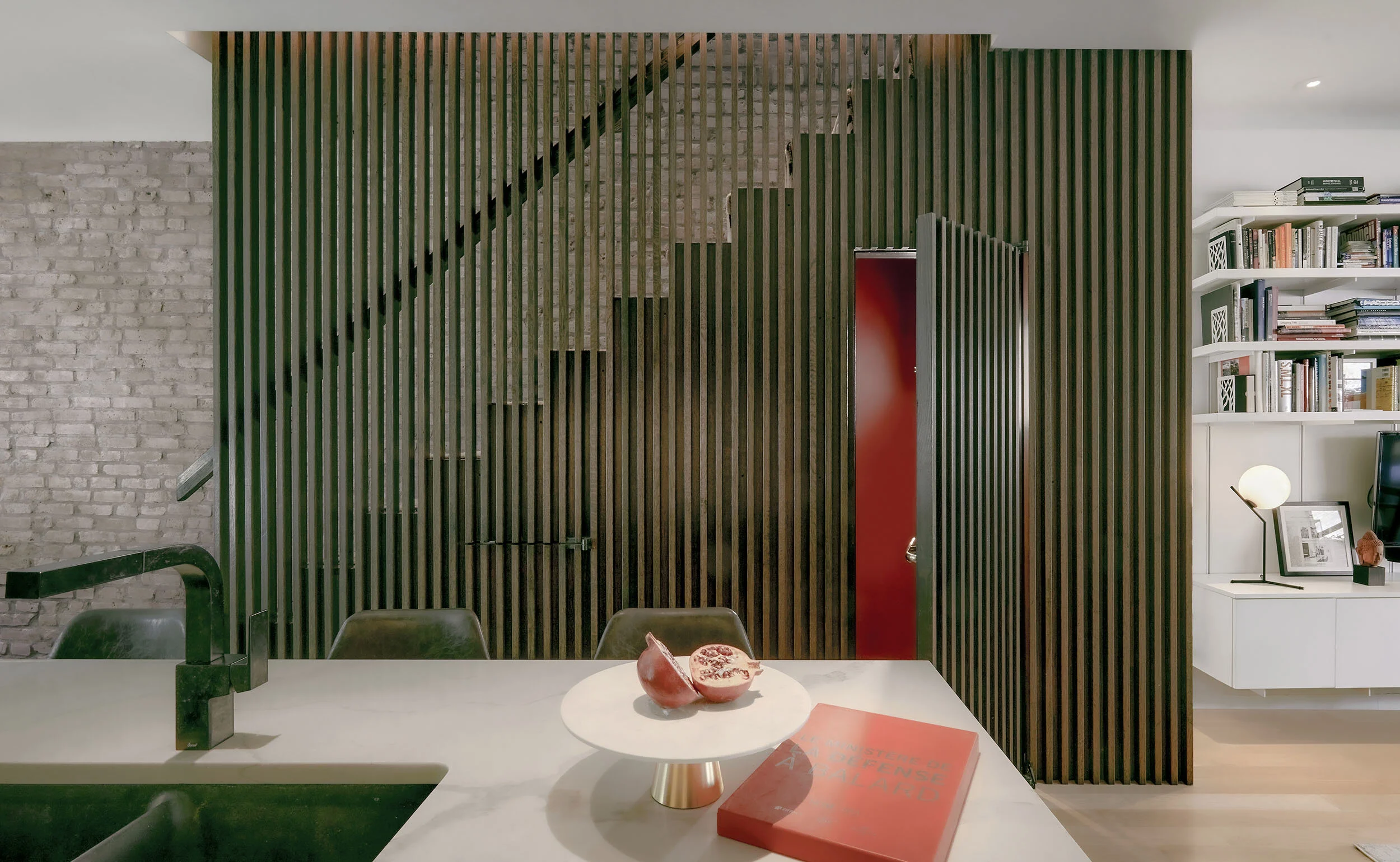 Modern interior with a slatted wood staircase, exposed brick wall, and a minimalistic table with a pomegranate and book. Open shelving with books and decorative items visible in the background.