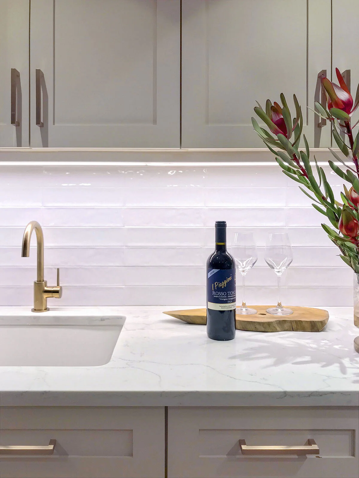 Modern kitchen counter with a marble surface, gold faucet, white subway tile backsplash, a wine bottle, two glasses on a wooden board, and a floral arrangement.