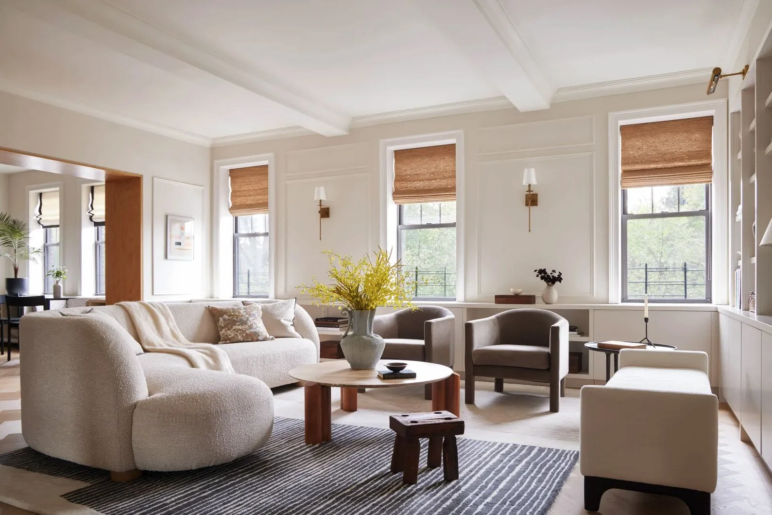 Modern living room with light-colored sofa, armchairs, and coffee table, decorated with yellow flowers and a rug, large windows with woven blinds, and neutral tones.