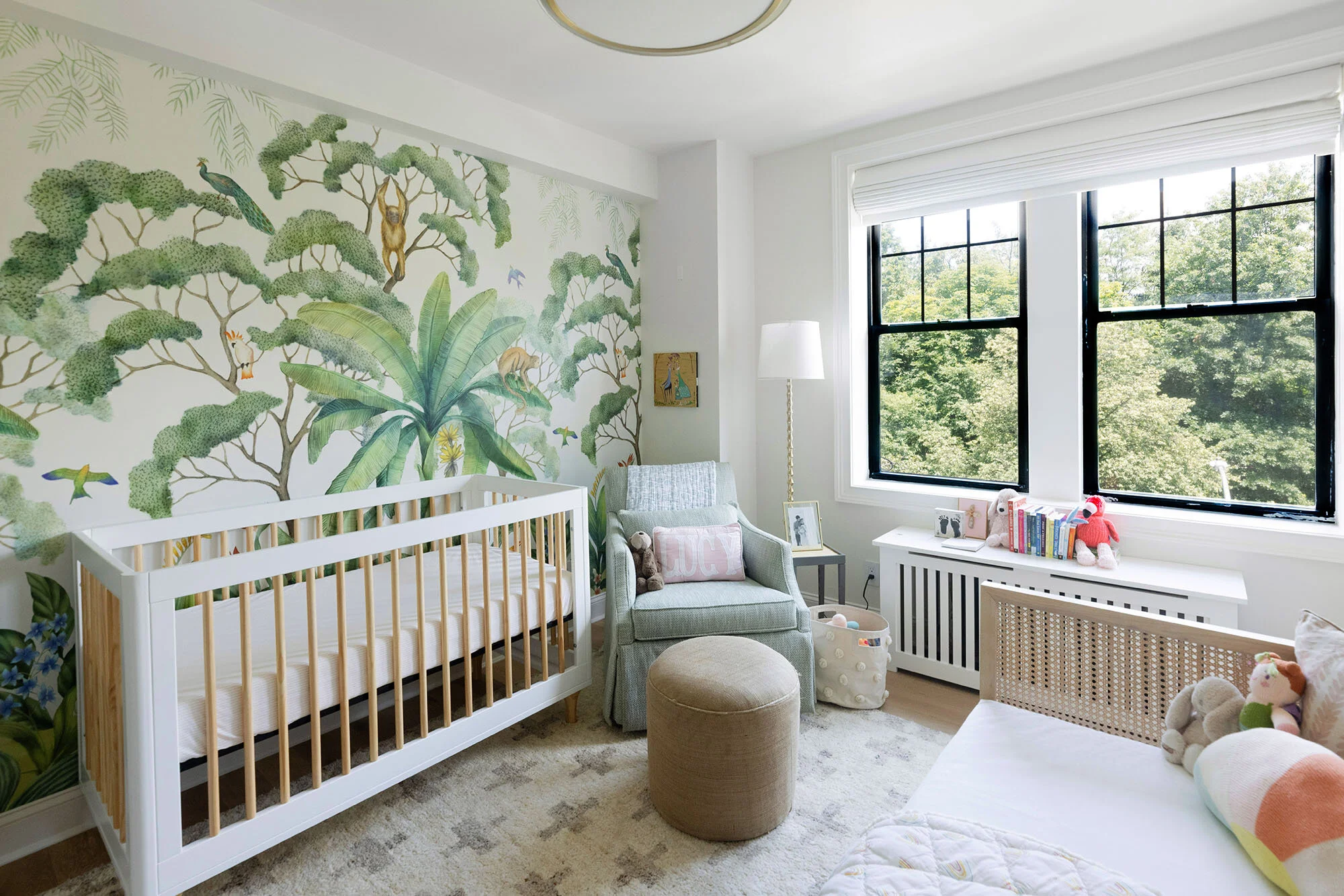 Nursery room with a jungle-themed mural, white crib, gray armchair, stuffed toys, books, and large windows with greenery outside.
