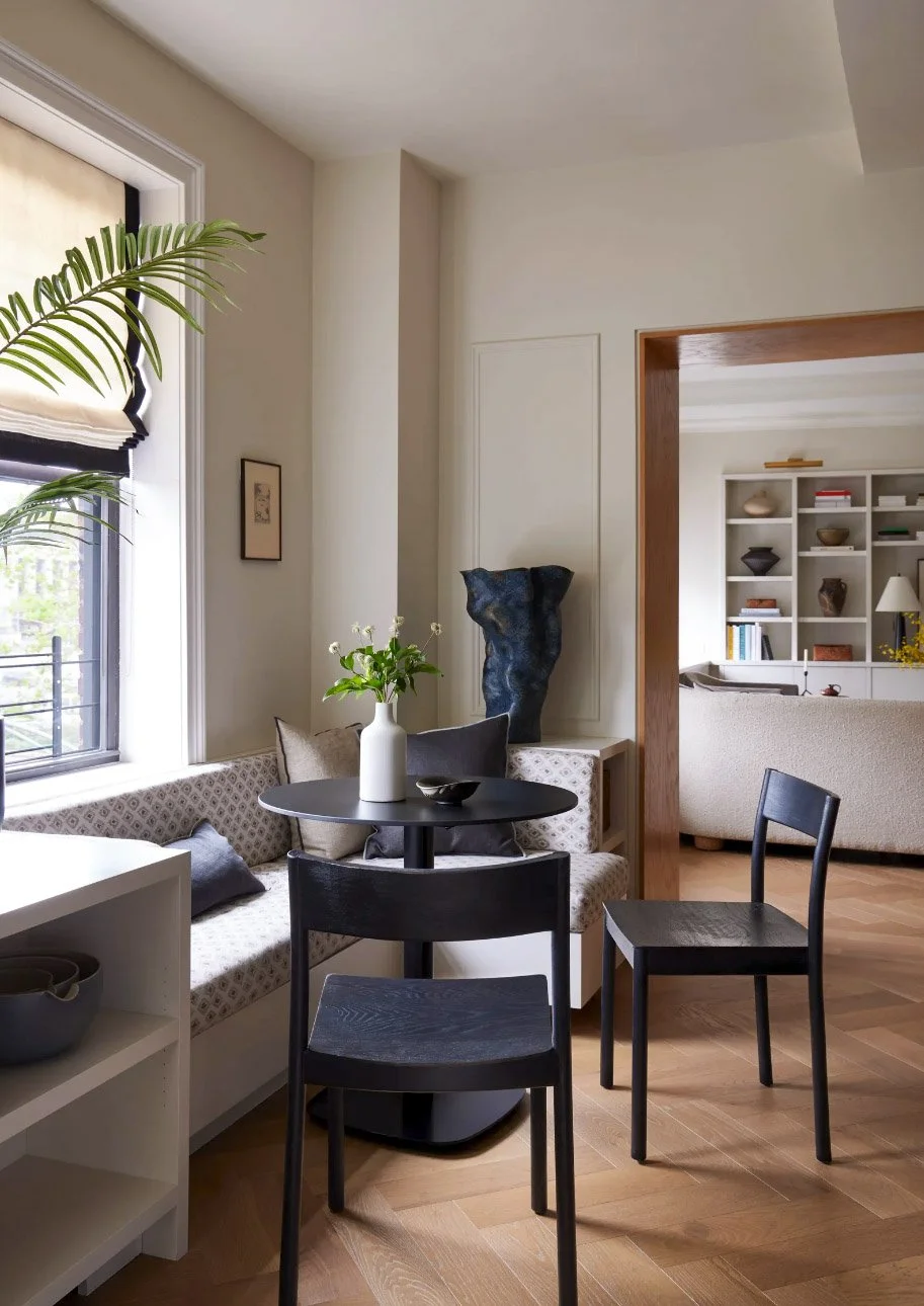 Modern dining nook with a black table, two chairs, a corner sofa, and a white vase with flowers. Nearby, there's a window with a beige shade, and in the background, a living room with a bookcase.