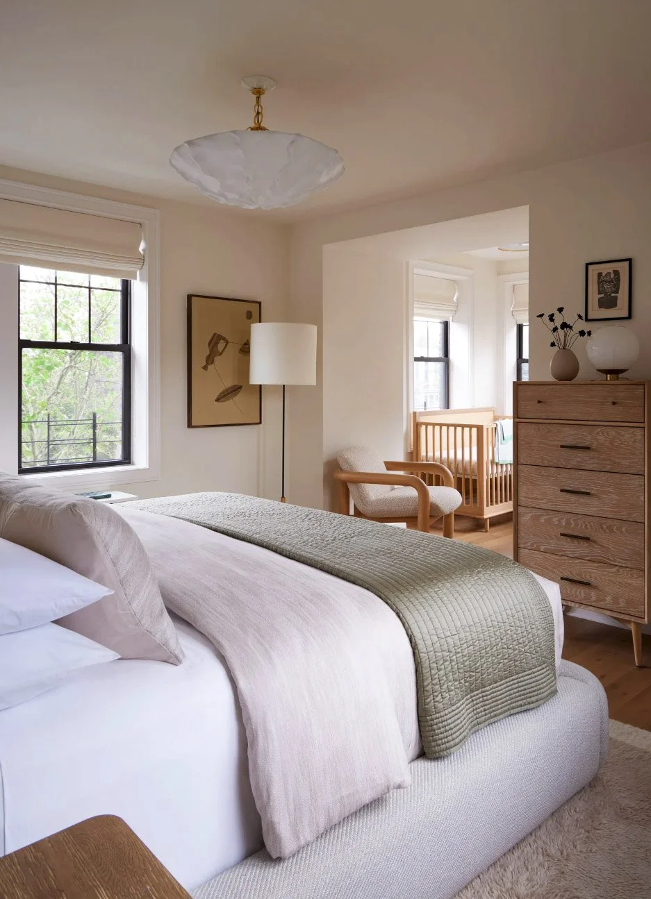Cozy bedroom interior with a neatly made bed featuring soft pillows and blankets, a wooden crib in the corner, a modern dresser, and a floor lamp. Natural light filters through large windows with Roman shades, enhancing the neutral color palette.