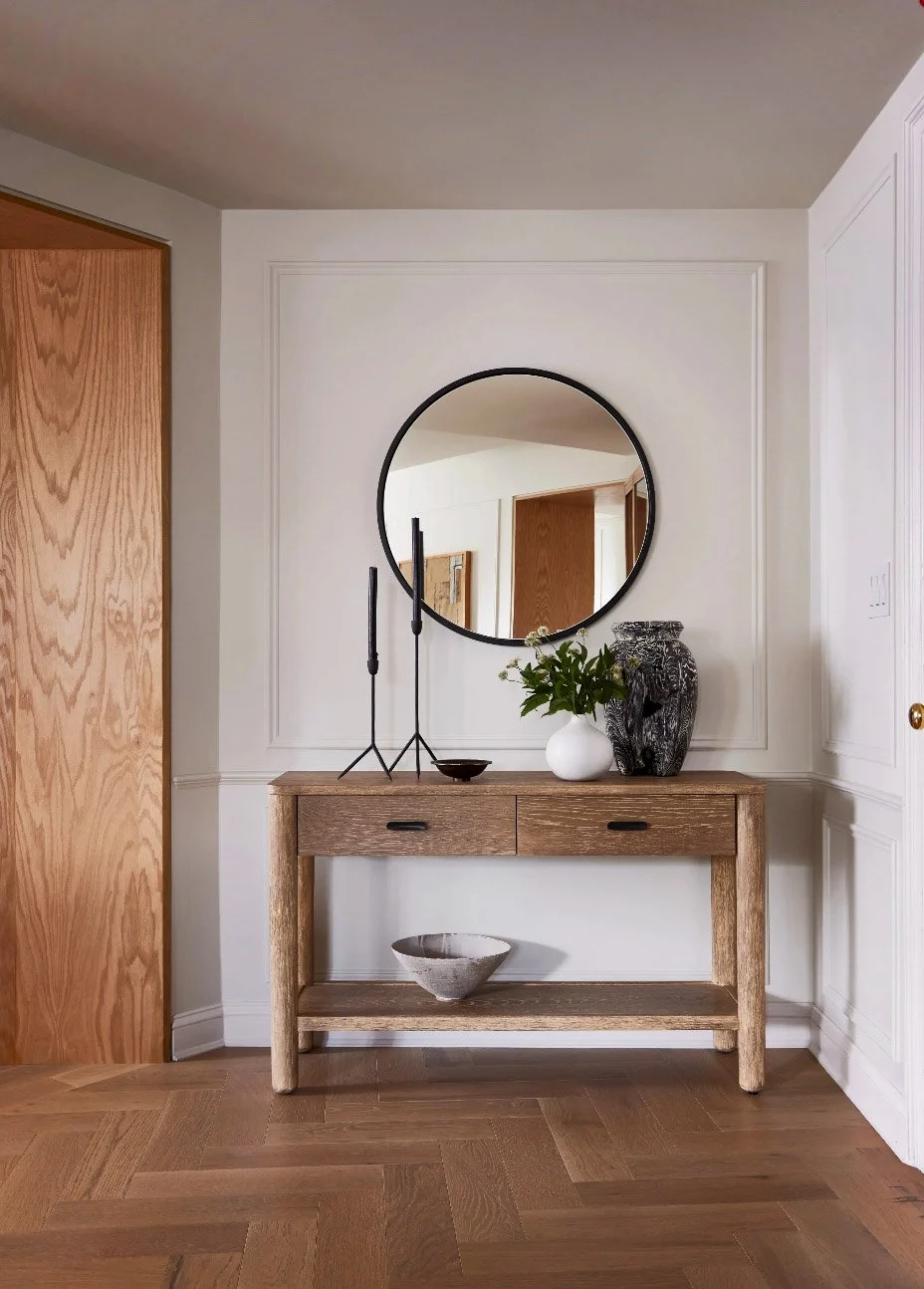 Stylish entryway with wooden console table, round mirror, tall black candlesticks, white vase with flowers, and decorative pottery, set against a white paneled wall.