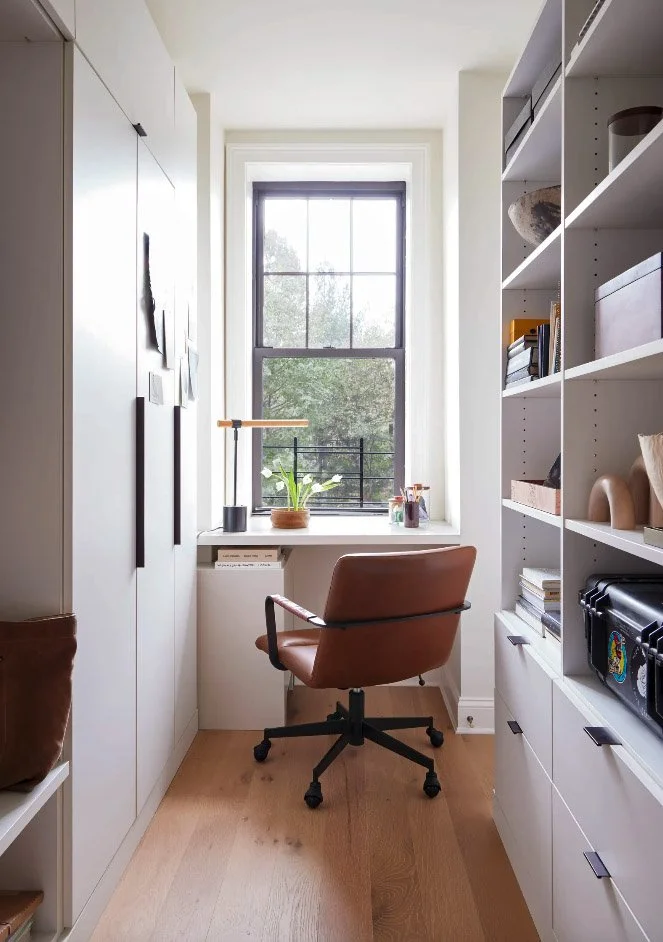 Small home office with a brown swivel chair, wooden desk, and shelving. A window with a potted plant and a lamp provides natural light. Floor is wooden, and shelves hold books and decorations.