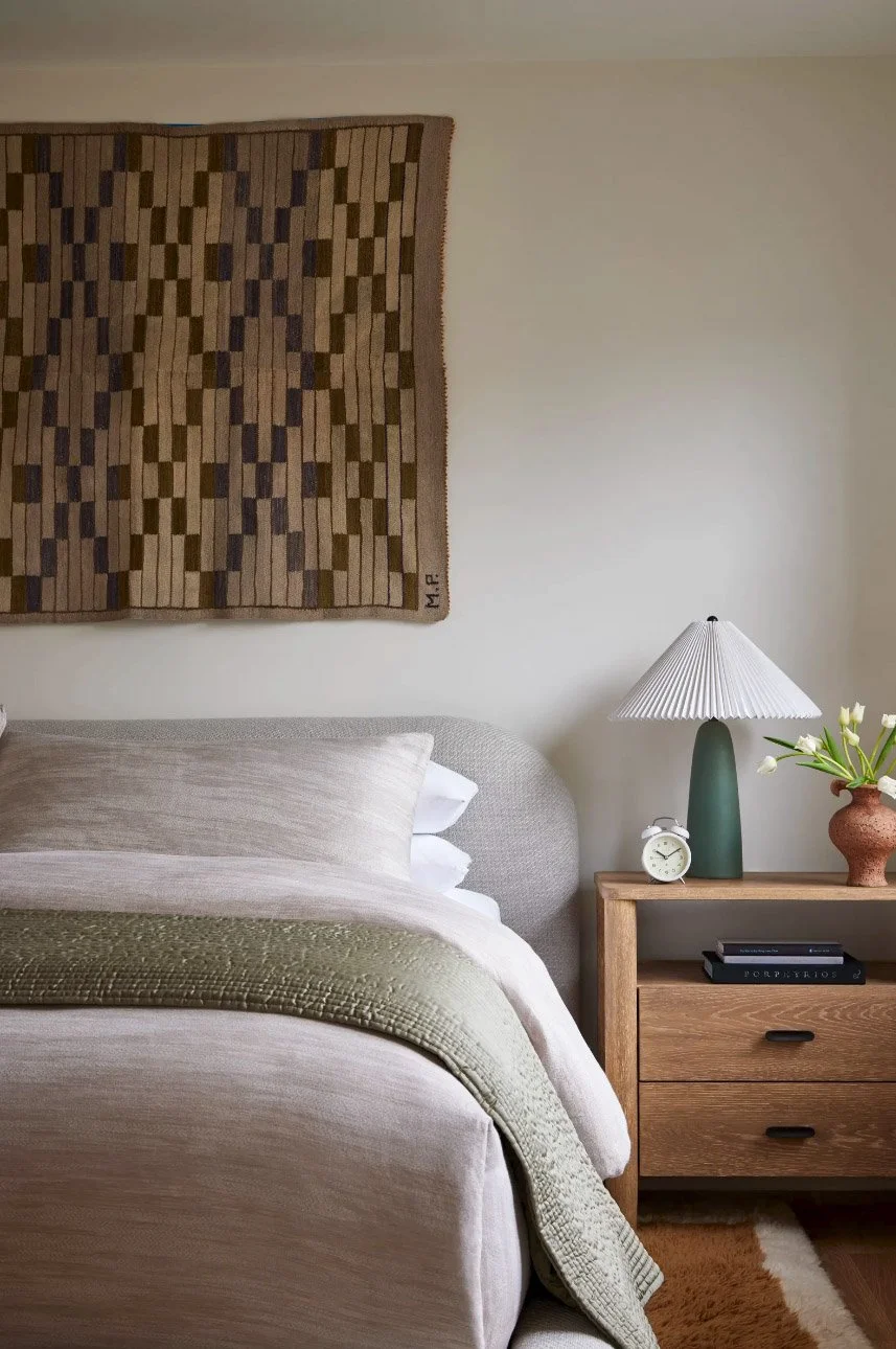 Cozy bedroom with neutral tones, featuring a bed with beige linens and a green throw, a wooden nightstand with a lamp, alarm clock, and decorative objects. A geometric wall hanging is above the bed.