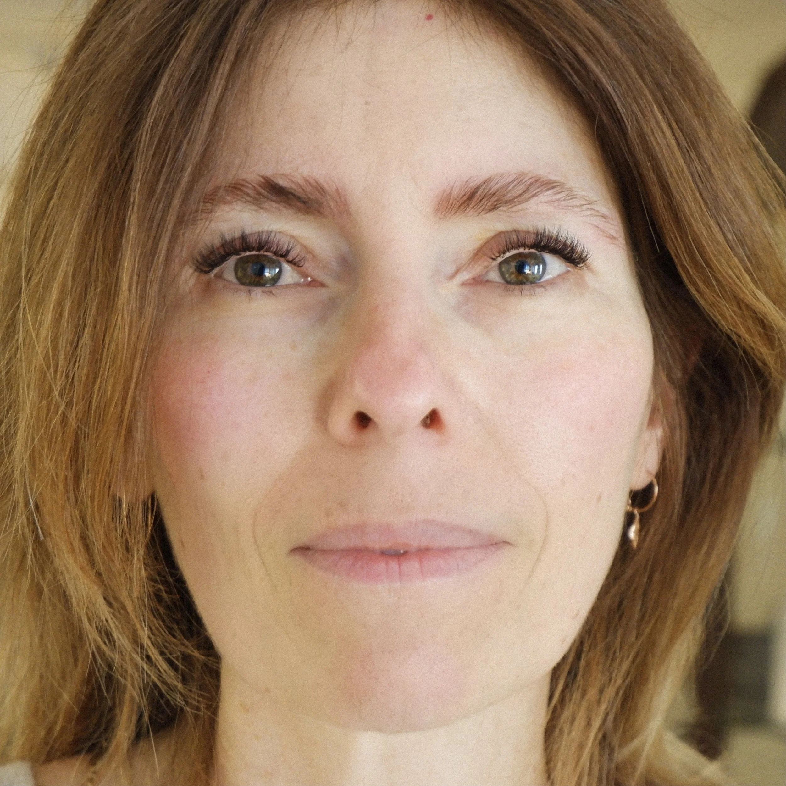 A woman's face with auburn hair and hazel eyes, wearing a gold earring. Natural lash extensions.