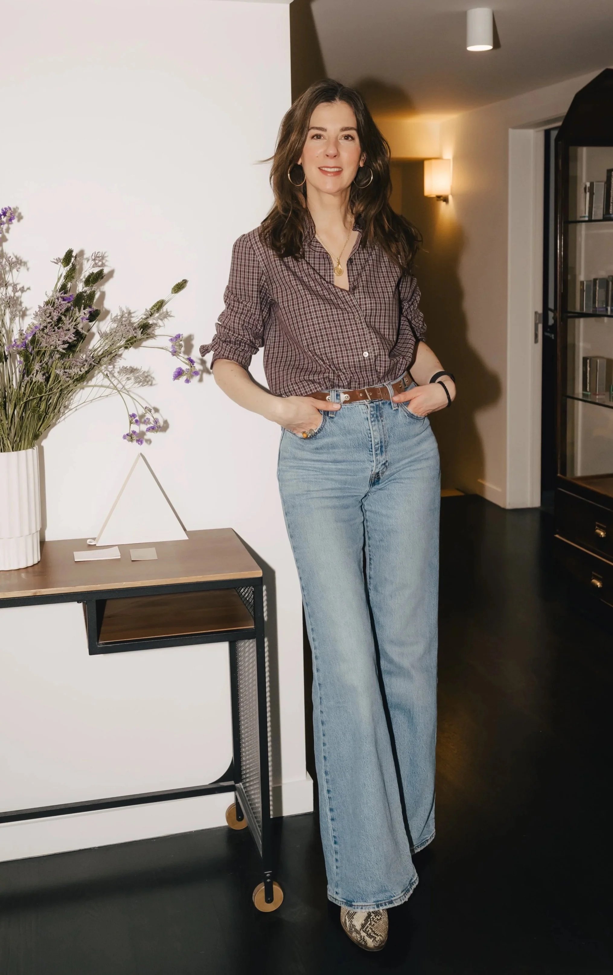 Une femme souriante avec des cheveux bruns, portant une chemise à carreaux, un jean à jambes larges, et des chaussures à motif serpent, se tient dans une pièce moderne avec une table basse, un vase de fleurs, et une bibliothèque en arrière-plan.