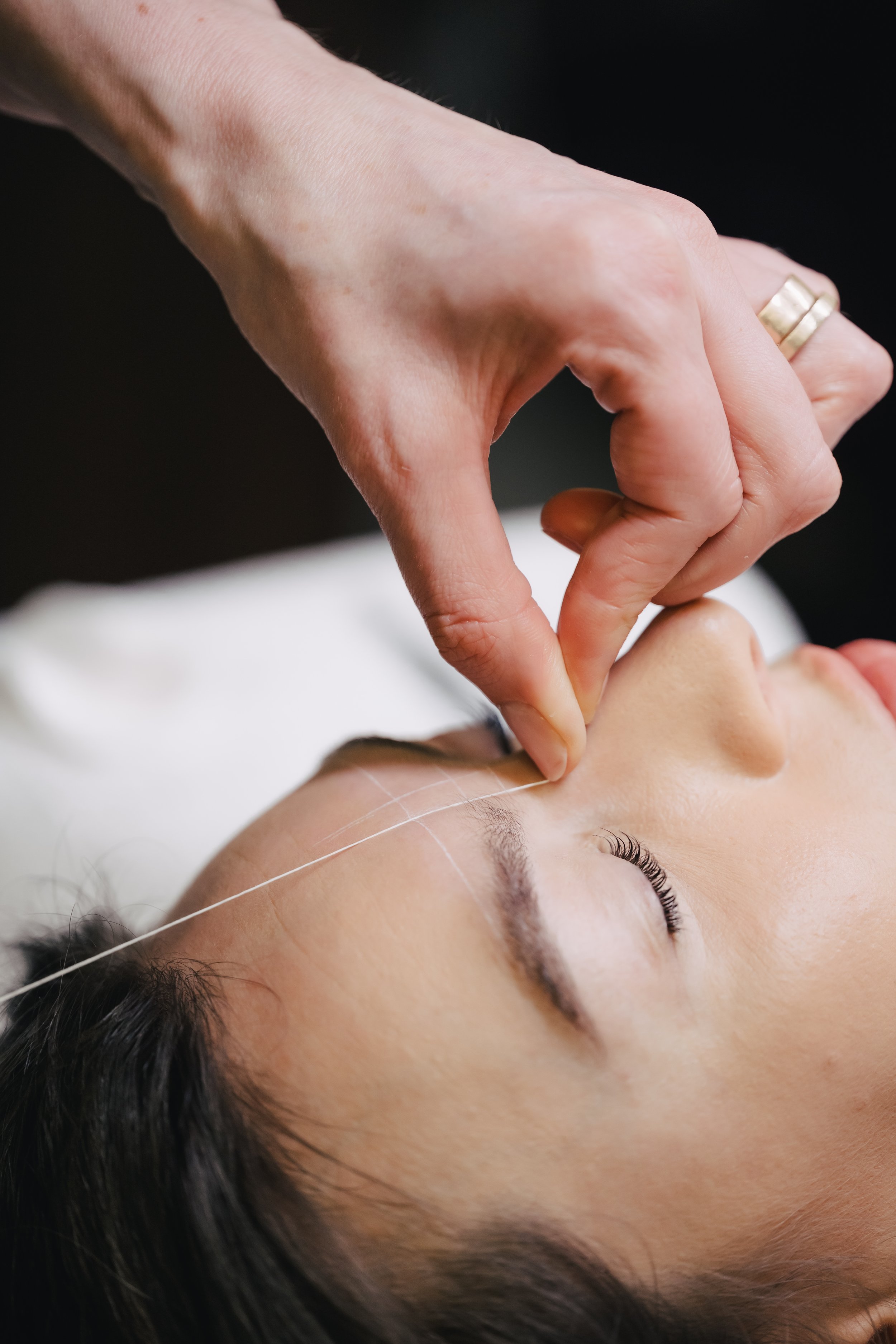 Une femme reçoit un soin esthétique ou une procédure de maquillage autour du contour des yeux, réalisée par une praticienne avec précision. Restructuration des sourcils et épilation