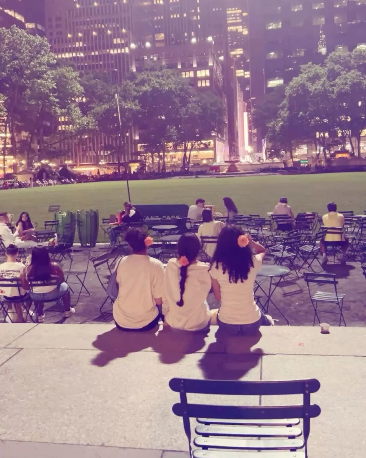 Trois femmes assises sur un rebord regardant un parc avec des chaises et des personnes assises ou se promenant. En arrière-plan, des bâtiments modernes et des arbres, avec un éclairage de nuit violet.
