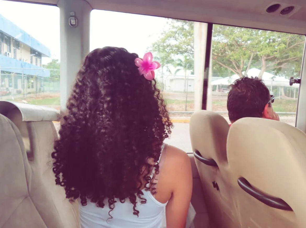 Une femme avec des cheveux bouclés portant une fleur rose dans les cheveux, assise dans un bus, regardant par la fenêtre.