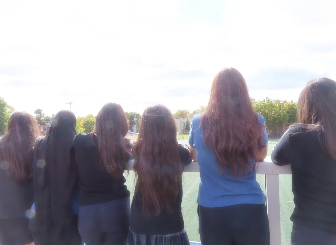 Cinq filles regardant par-dessus une barrière sur un terrain de sport extérieur, avec un ciel partiellement nuageux et des arbres en arrière-plan.