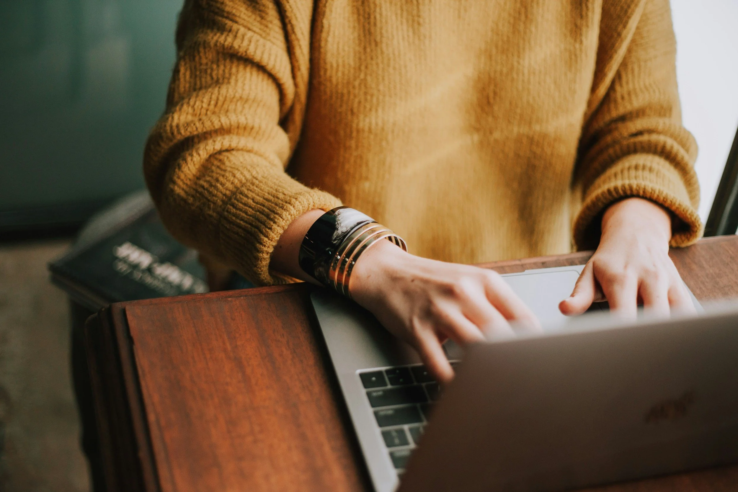 A person wearing a tan sweater typing on a laptop at a wooden table.