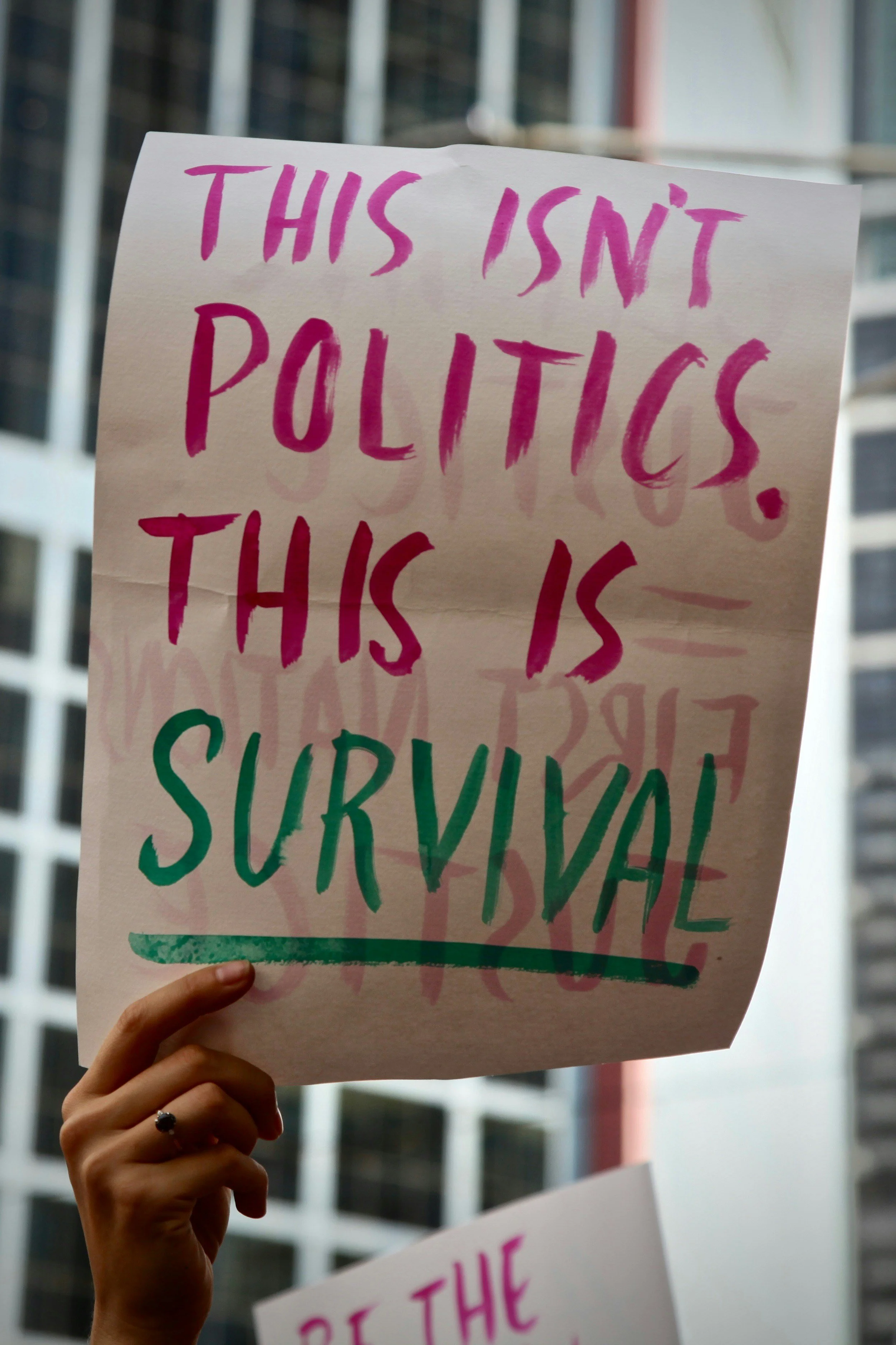 A protestor holding a sign that reads 'This isn't politics. This is survival.' in pink and green letters, with buildings in the background.