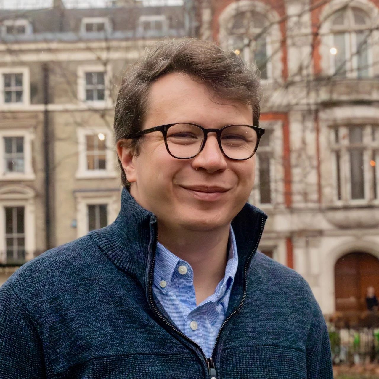 Young man with glasses and dark hair, smiling, wearing a white button-up shirt, standing against a plain light-colored wall.