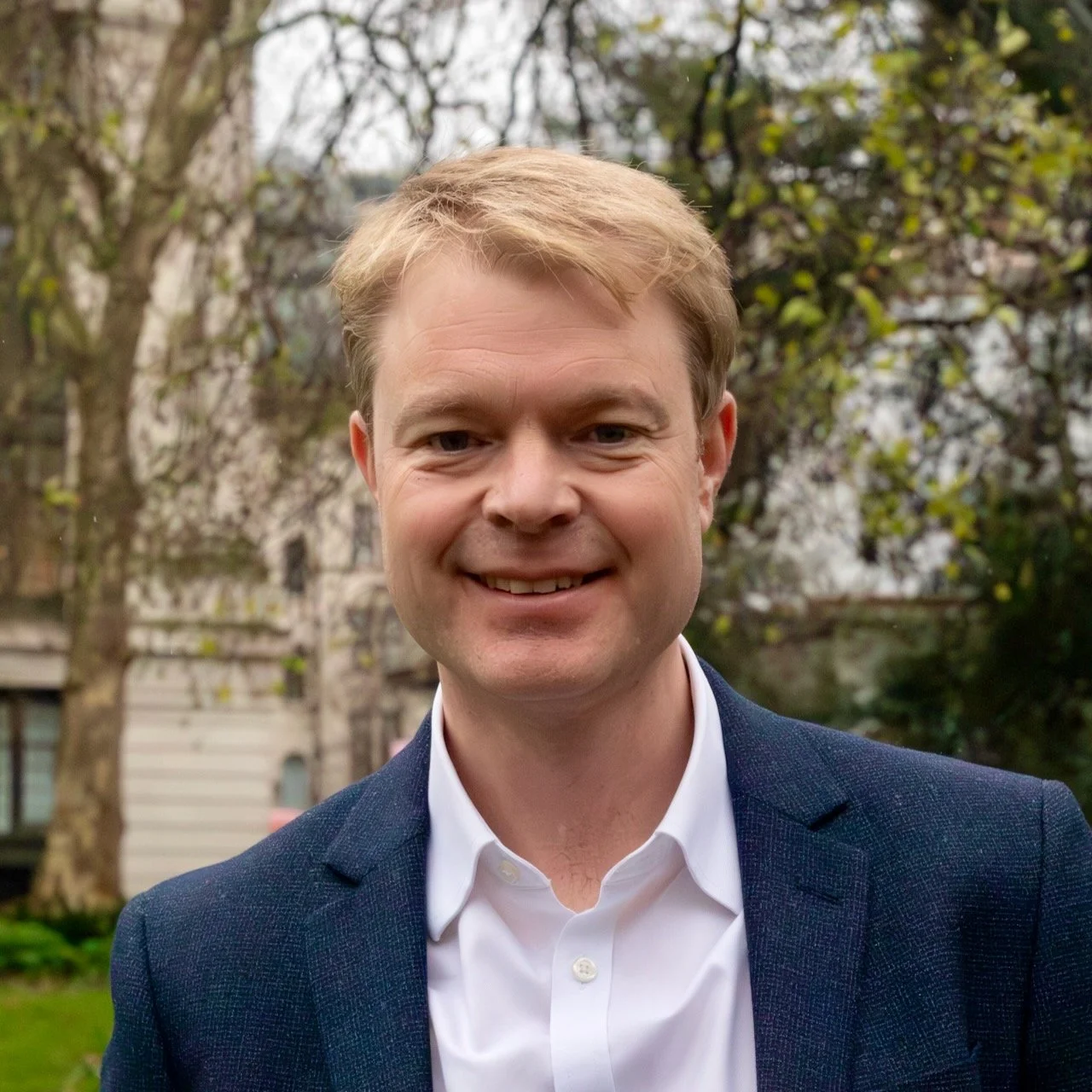 A young man with light skin and reddish-blonde hair standing in front of a plain, light-colored wall, wearing a light blue button-up shirt.