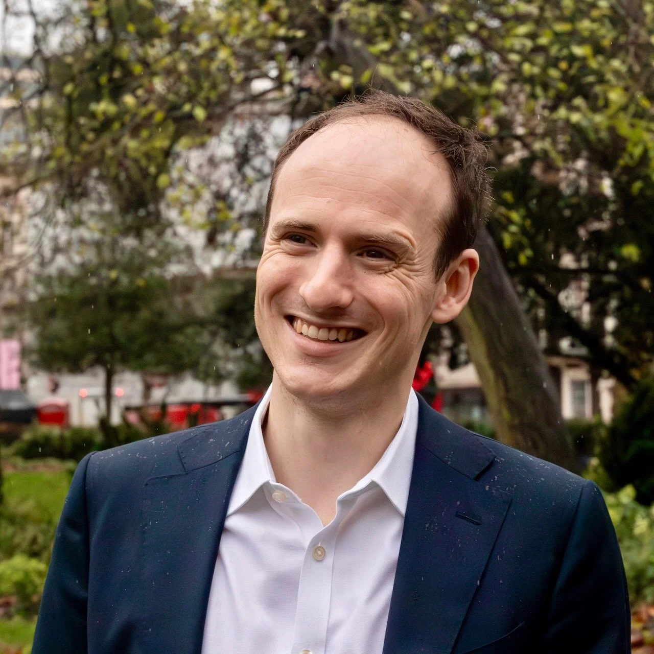 A man with short dark hair and fair skin standing against a plain beige wall, wearing a white collared dress shirt.