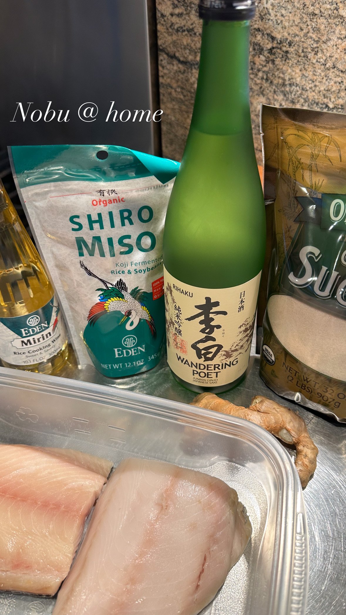 A bottle of Junmai sake labeled 'Wandering Poet' beside a bag of organic shiro miso, ginger root, some raw fish fillets, and other cooking ingredients on a kitchen counter.