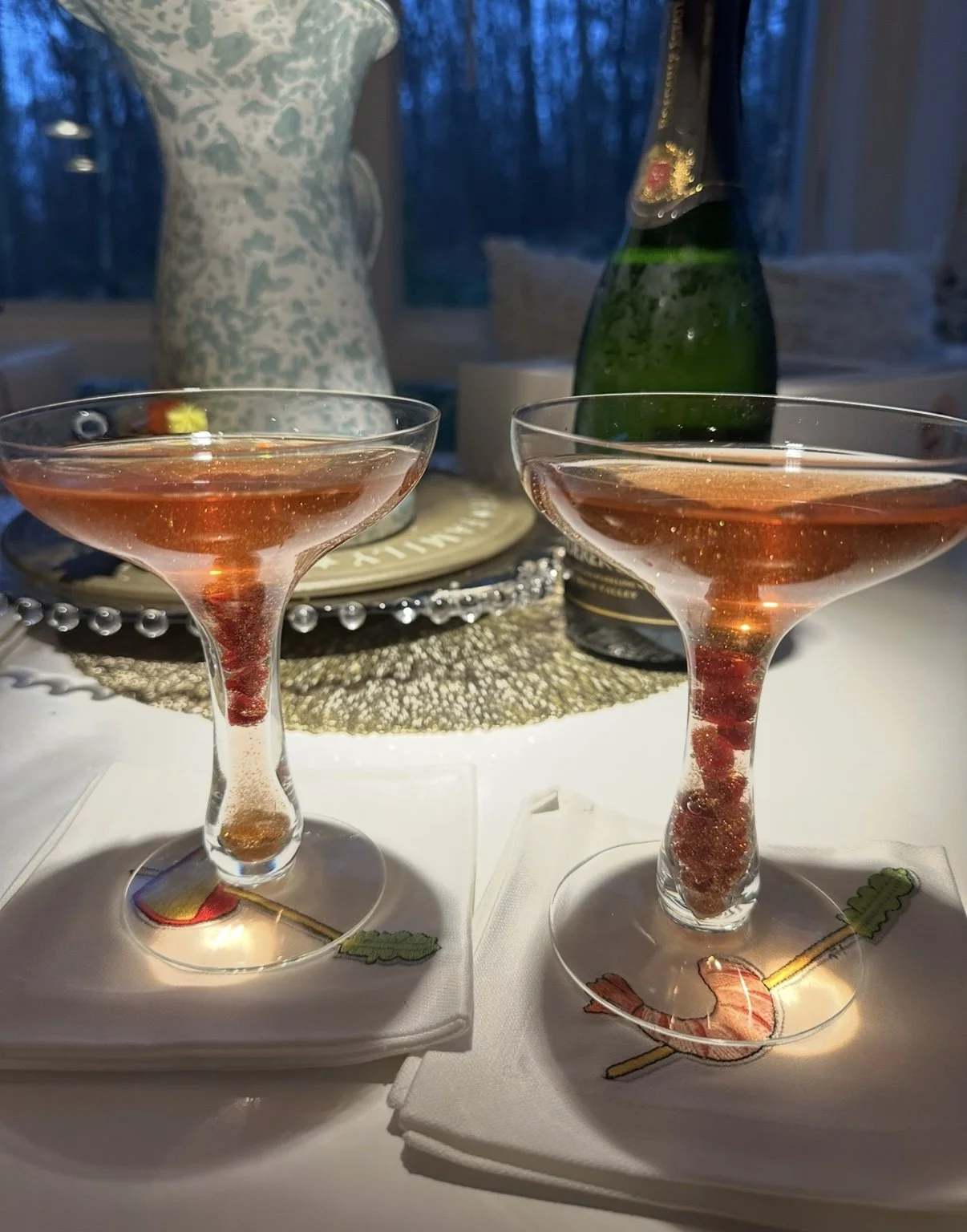 Two glasses of pink champagne on napkins with colorful cocktail stickers, a bottle of sparkling wine in the background, and a decorative vase on a table.